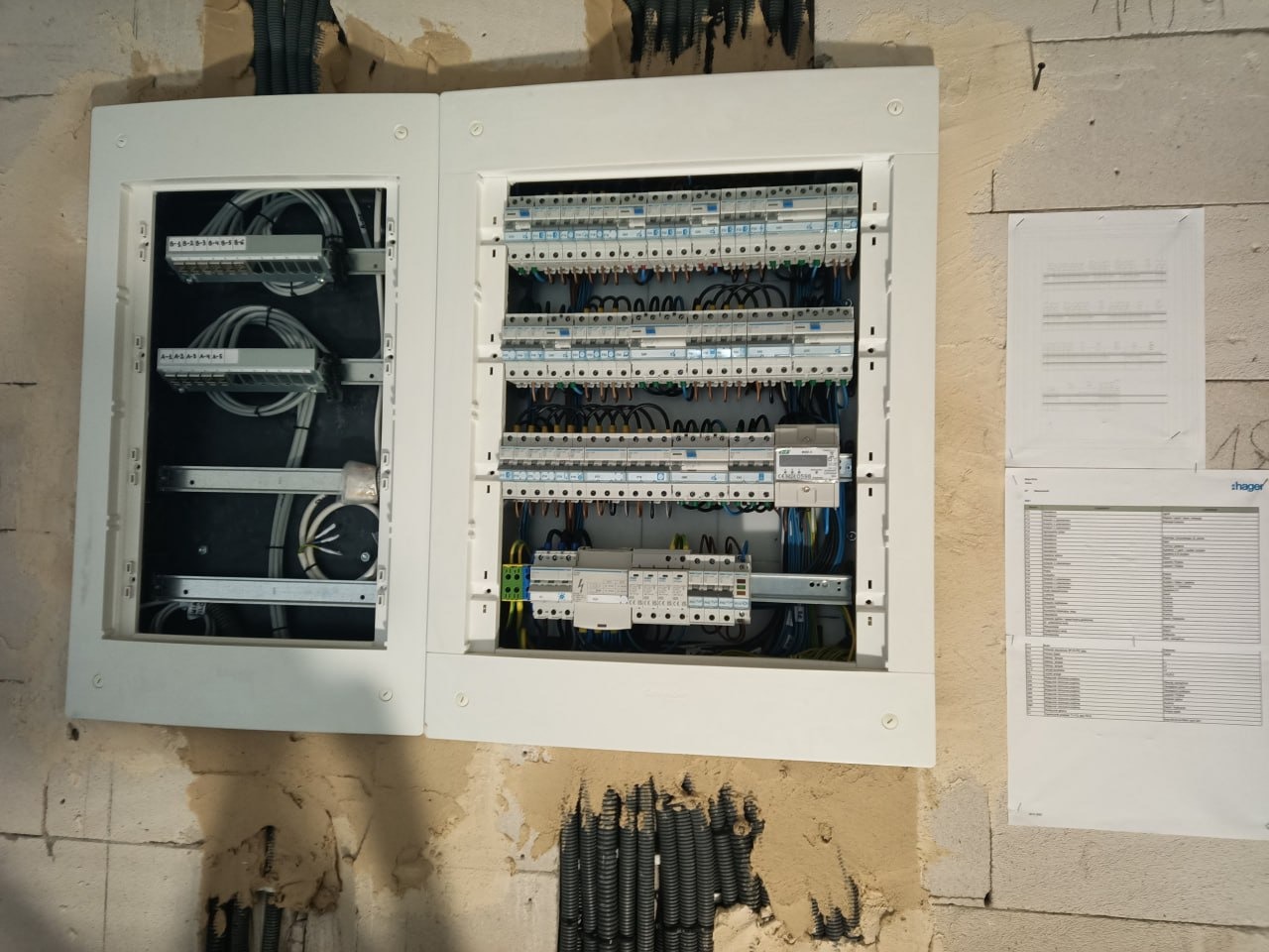 Otwarta, biała skrzynka rozdzielcza z widocznymi kablami i bezpiecznikami, zamontowana na surowej ścianie. Obok dokumentacja techniczna. Widoczne przewody w ścianie.