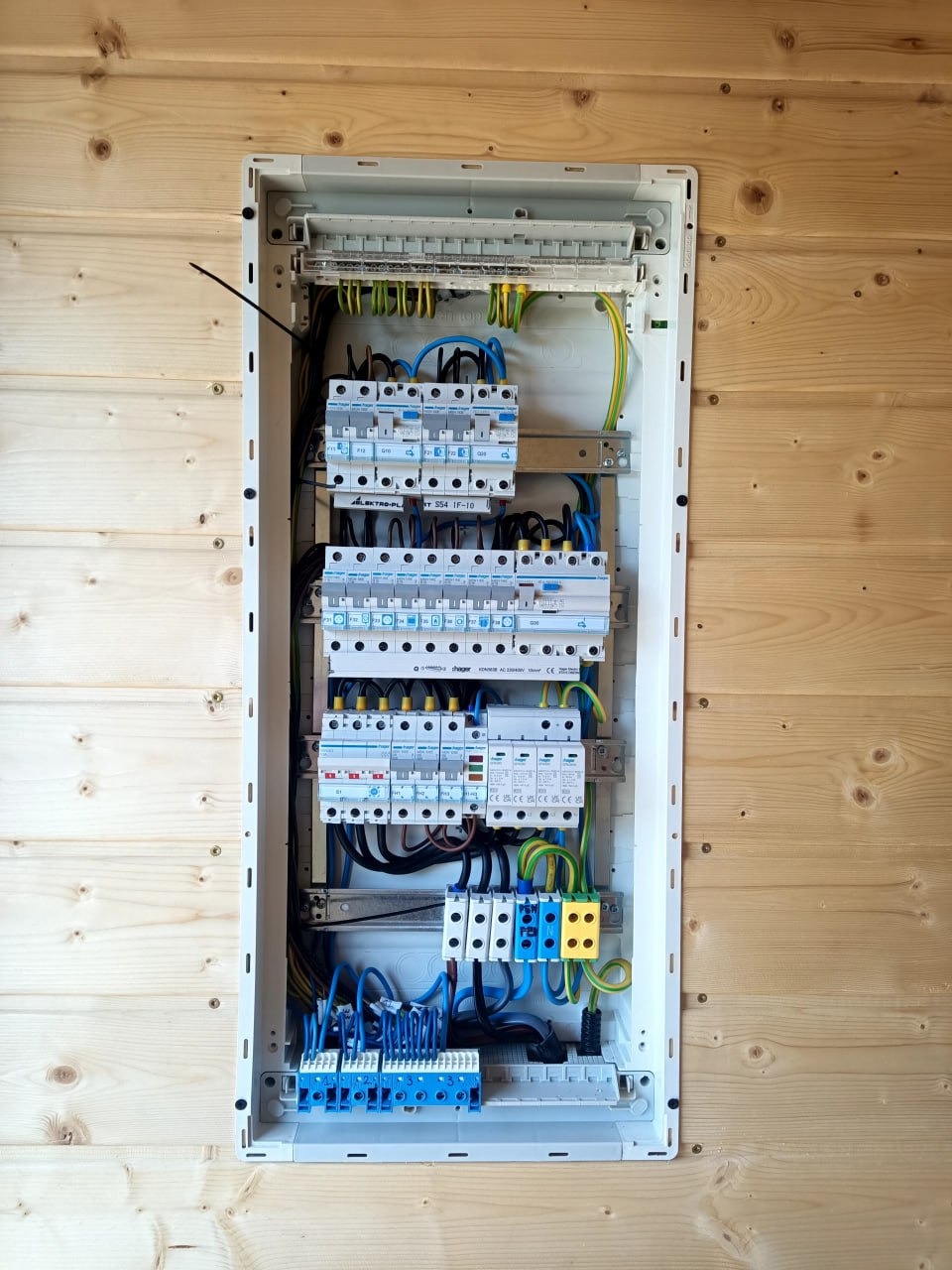 Rozdzielnica elektryczna Hager w obudowie, zamontowana na ścianie z boazerii. Widoczne zabezpieczenia, przewody w kolorach: niebieskim, czarnym, żółto-zielonym.