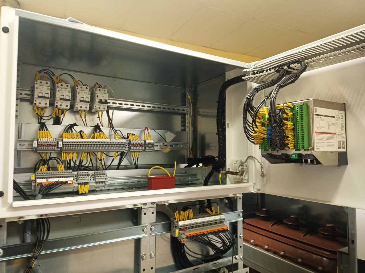 Otwarta szafa sterownicza z precyzyjnie ułożonymi przewodami oznaczonymi żółtymi etykietami, widoczne zabezpieczenia i listwy zaciskowe.