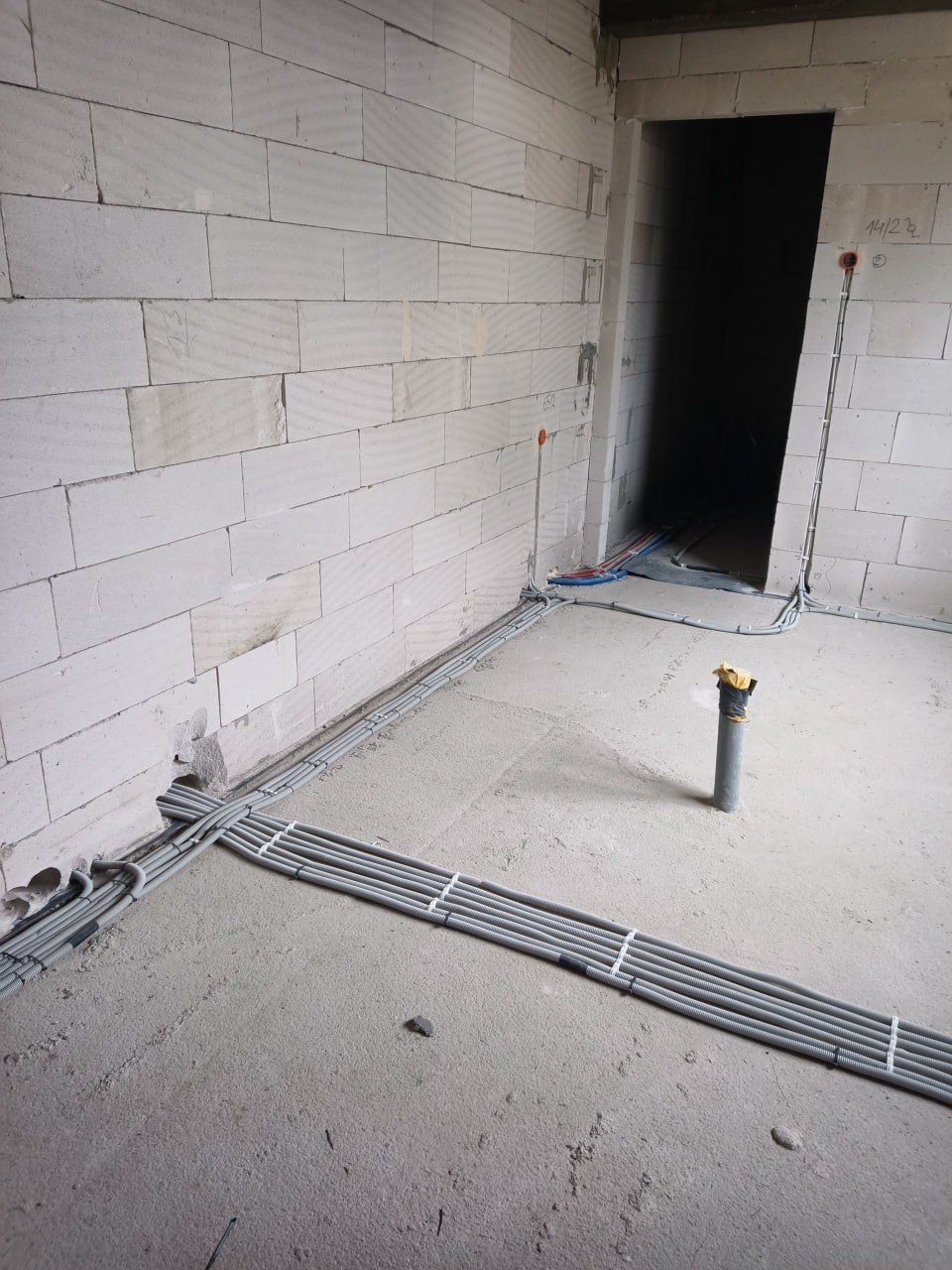 Szare rury w peszlu ułożone na surowym betonie podłogi w nowym budynku z białego pustaka. Część instalacji poprowadzona po ścianie. Widoczne otwory na gniazdka.