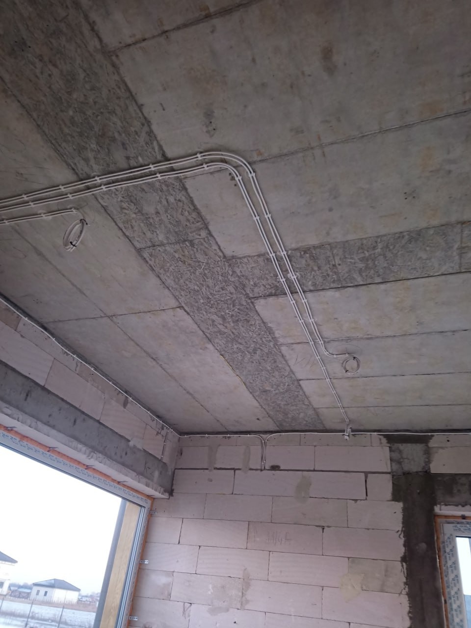 Białe kable elektryczne prowadzone po suficie z betonu i płyt OSB w surowym wnętrzu budynku w trakcie budowy. Widoczne okno i ściany z białych bloczków.