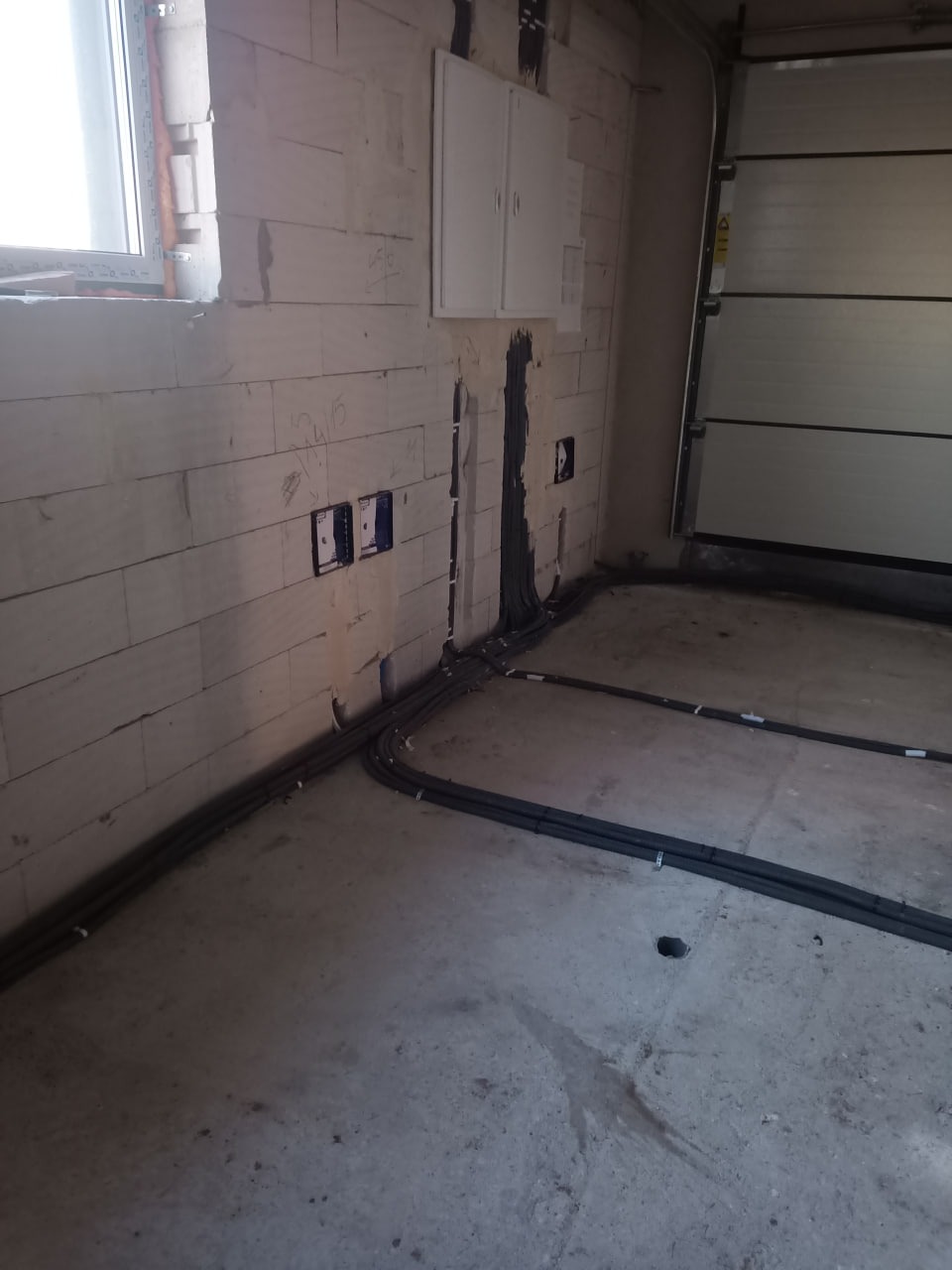 Szare kable w peszlu ułożone wzdłuż ściany z białego bloczka, widoczne puszki instalacyjne i skrzynka rozdzielcza. Garaż w tle. Surowy stan.