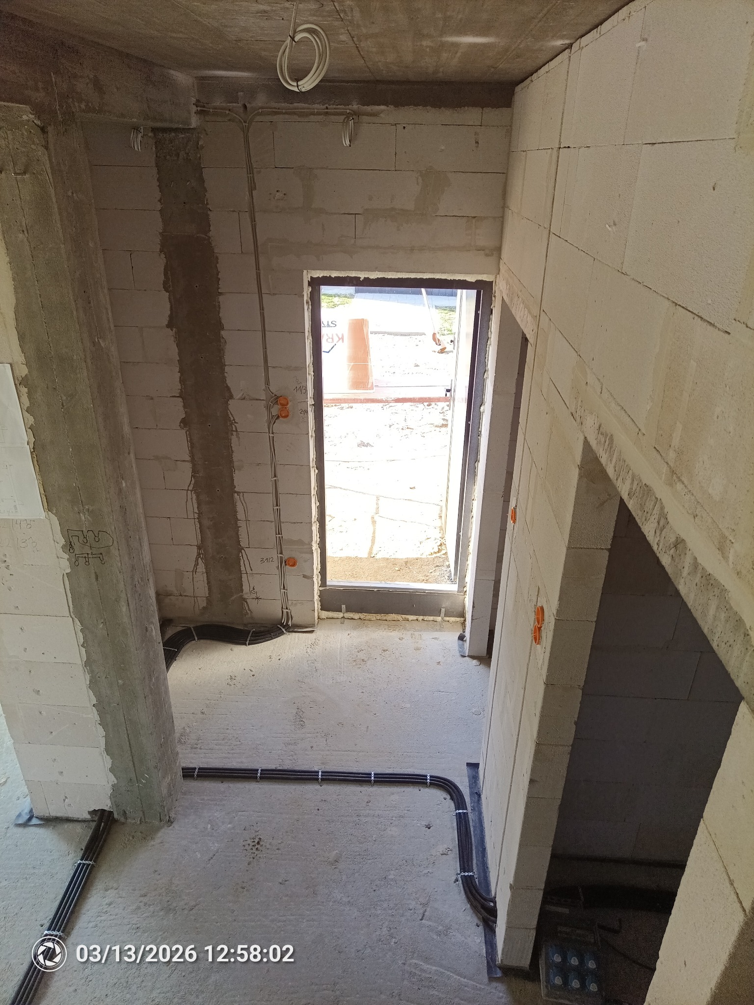 Surowy stan wnętrza z widocznymi przewodami elektrycznymi ułożonymi wzdłuż podłogi i ścian, pomarańczowe puszki elektryczne na ścianach, budynek w trakcie budowy.