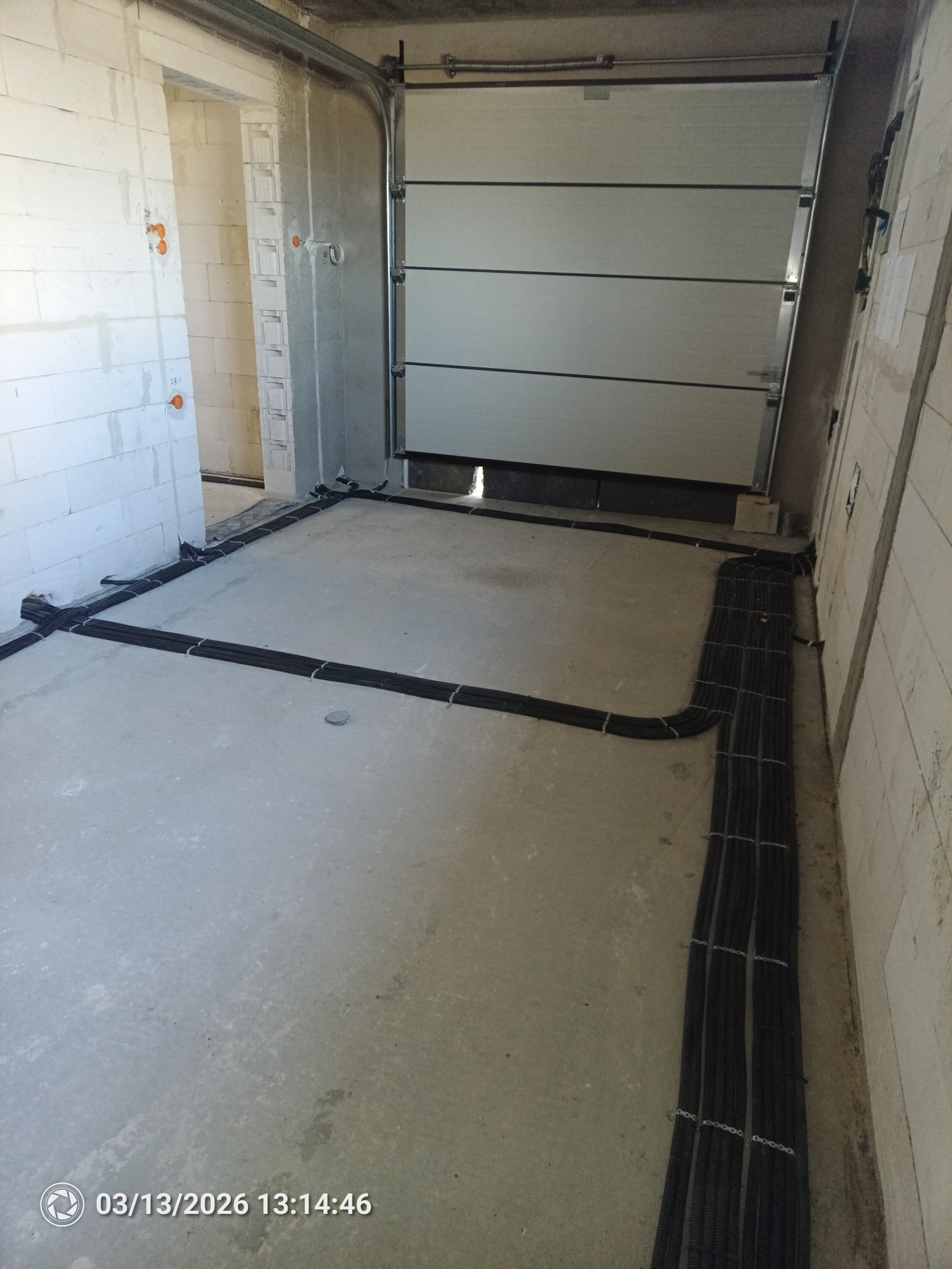 Kable elektryczne ułożone na podłodze w surowym garażu przed wylaniem posadzki. Widoczne ściany z białego bloczka i brama garażowa segmentowa.