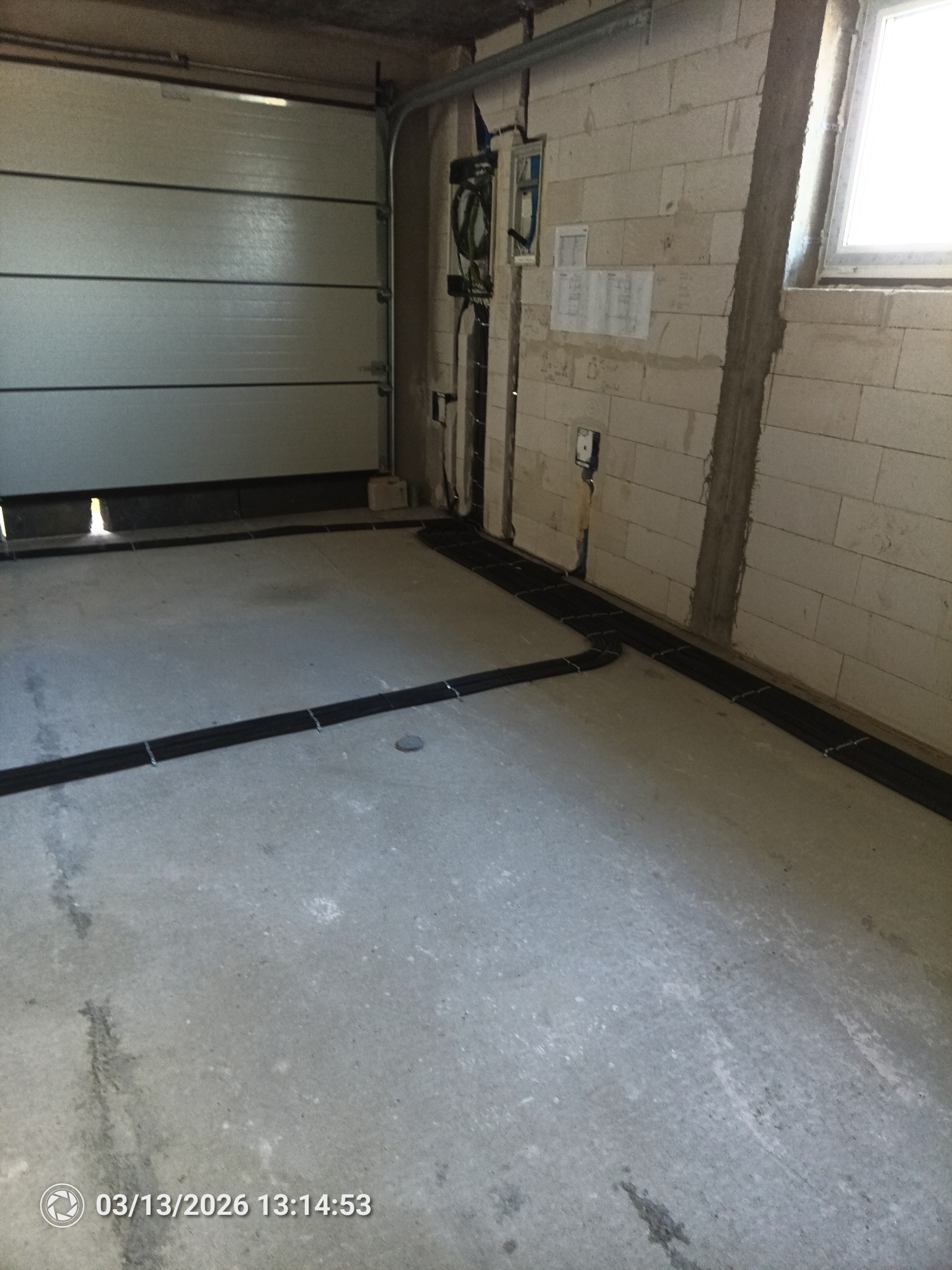 Układanie przewodów w peszlu na podłodze garażu, przygotowanie do zalania posadzki. Widoczne kable prowadzone wzdłuż ścian, surowy stan budynku.