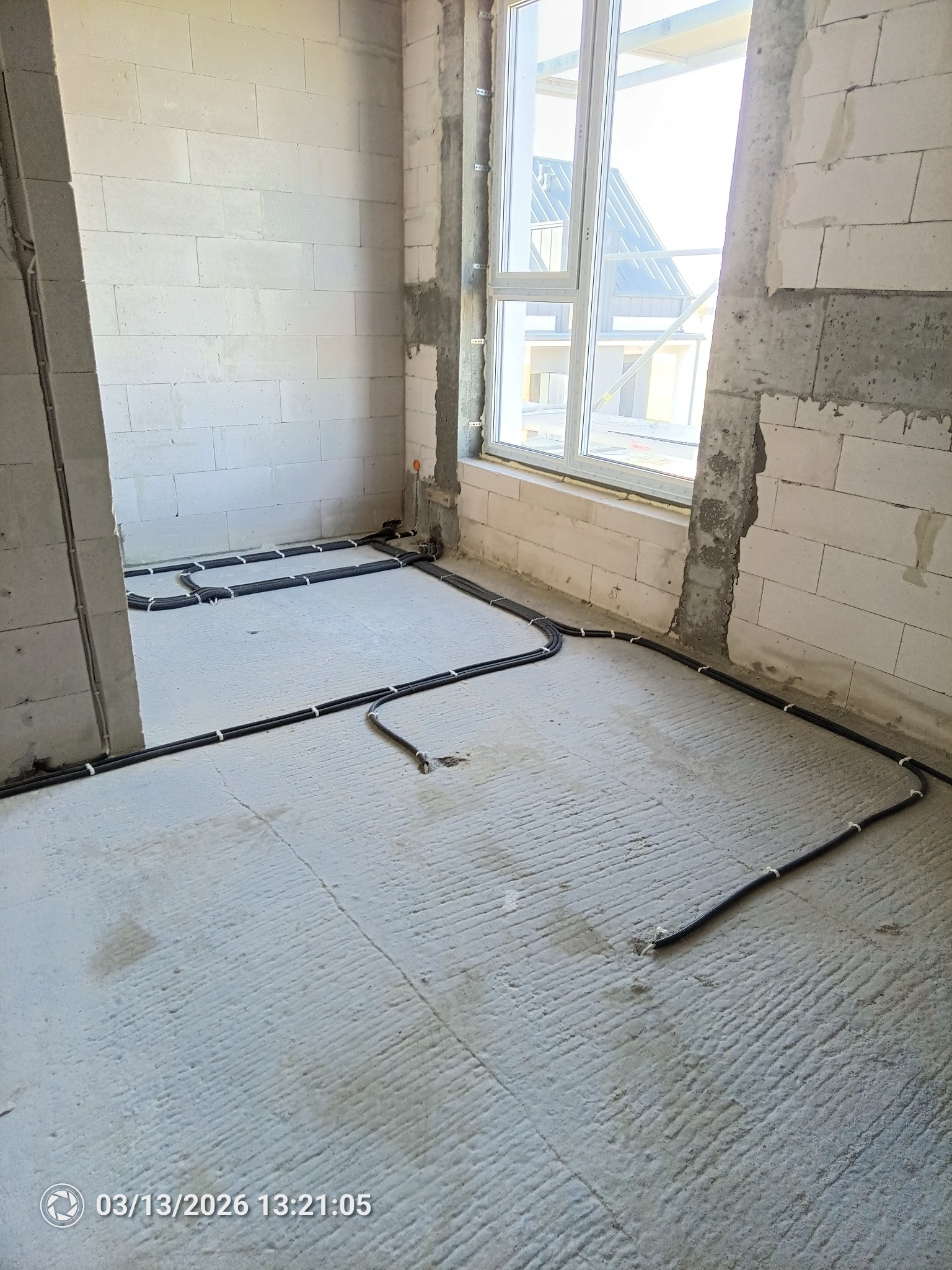 Wnętrze w trakcie budowy z widocznymi przewodami w peszlach ułożonymi na podłodze, przygotowanymi do zalania w posadzce. Okno w tle doświetla przestrzeń.