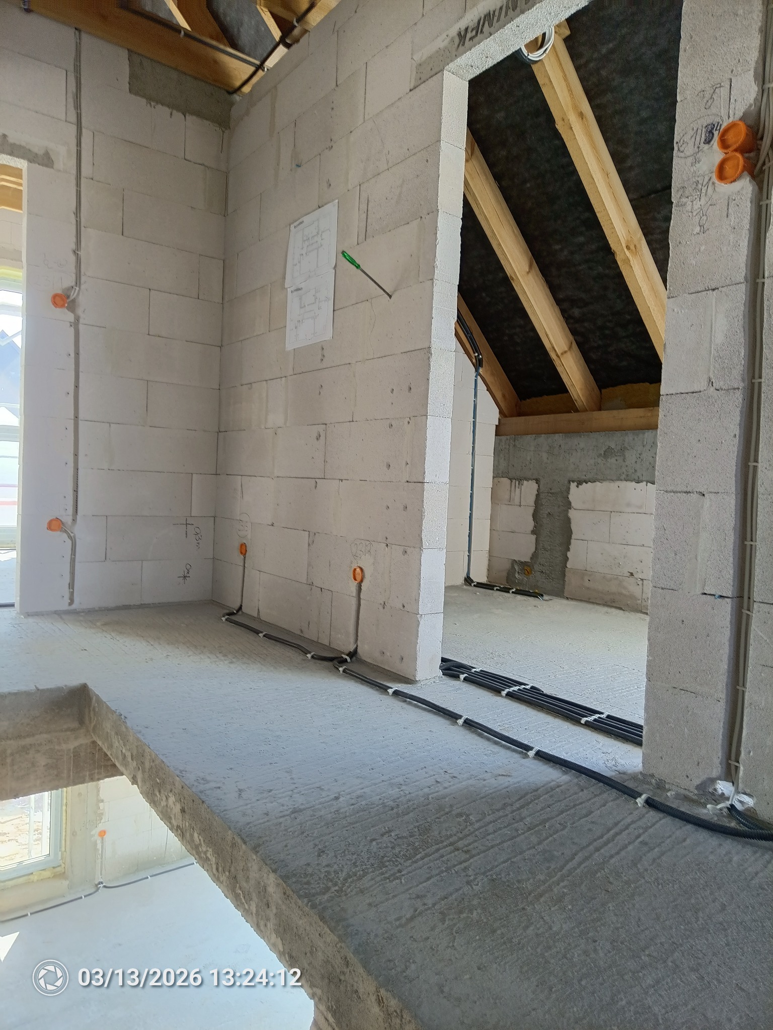 Wnętrze w stanie surowym z widocznymi przewodami elektrycznymi ułożonymi na podłodze i wyprowadzonymi ze ścian. Widoczne puszki instalacyjne i plan budynku na ścianie.