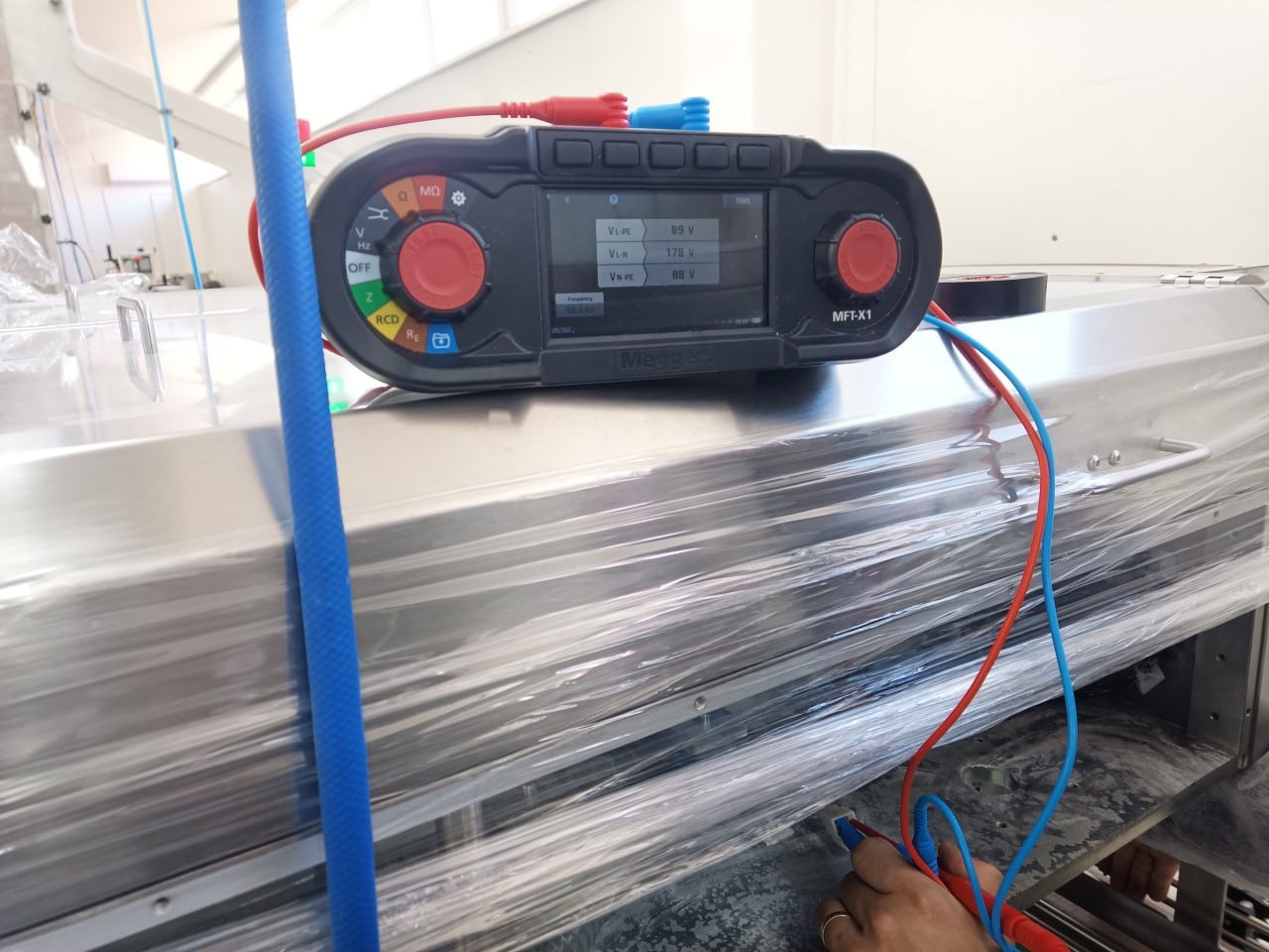 Miernik wielofunkcyjny Megger MFT-X1 z podłączonymi przewodami pomiarowymi, wyświetlający wyniki pomiarów napięcia. Urządzenie leży na metalowej powierzchni.