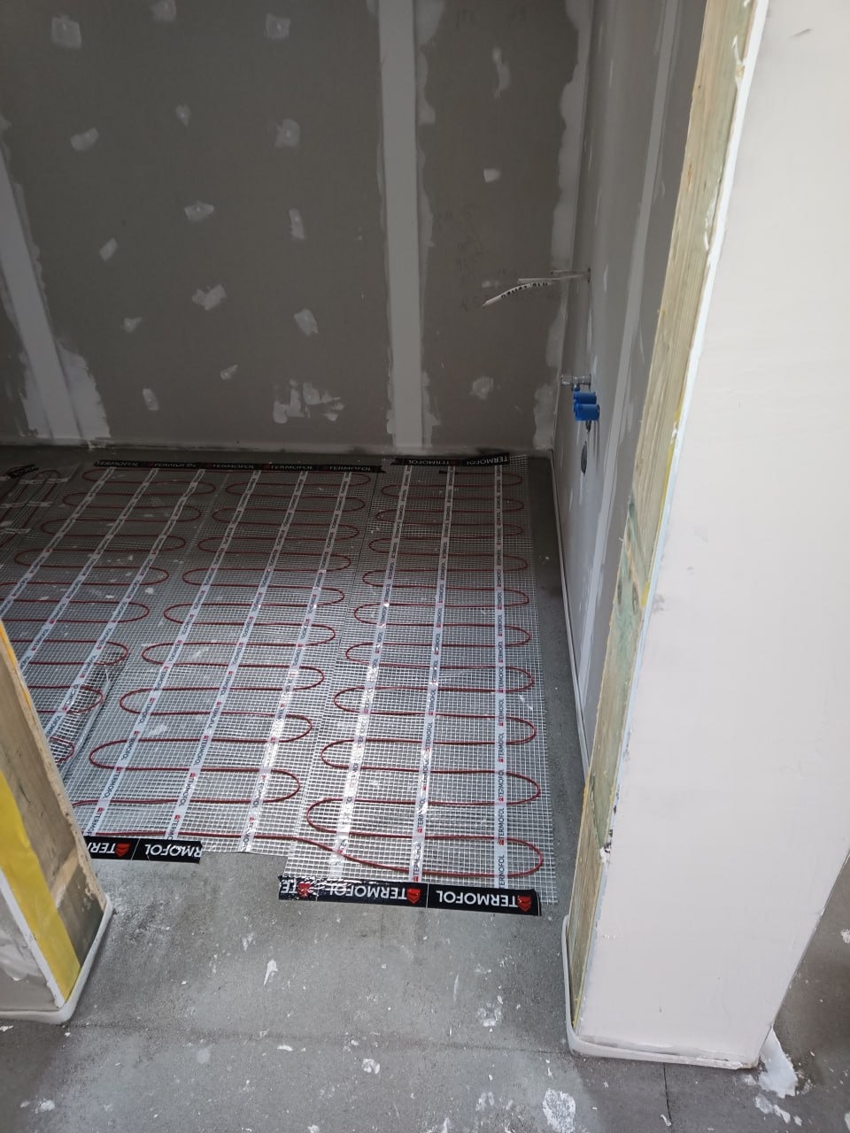 Elektryczne ogrzewanie podłogowe, mata grzewcza Termofol TE, czerwone kable na siatce, widoczne puszki elektryczne w surowym wnętrzu przed wylewką.