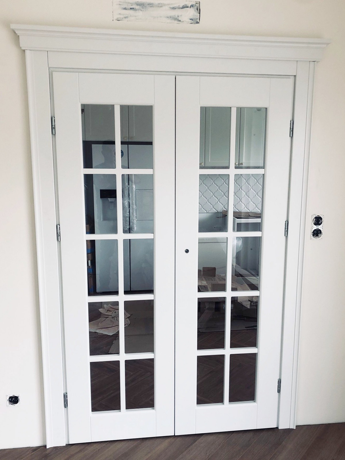 Białe, przeszklone drzwi dwuskrzydłowe z ozdobną ramą, zamontowane w jasnym wnętrzu. Widoczne gniazdka elektryczne i fragment podłogi.