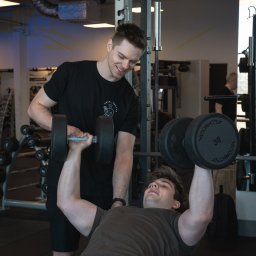 Trener Personalny Arek Warpechowski - Mężczyzna ćwiczy wyciskanie sztangielek pod okiem uśmiechniętego instruktora w siłowni. Widoczne regały z obciążeniami i sprzęt do ćwiczeń.