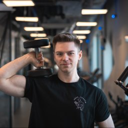 Trener Personalny Arek Warpechowski - Młody mężczyzna w czarnej koszulce z logotypem, trzymający hantlę na ramieniu w nowoczesnej siłowni z niebieskimi akcentami świetlnymi.