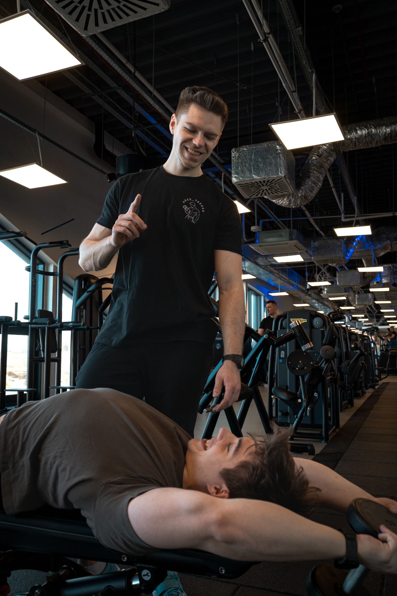 Uśmiechnięty mężczyzna w czarnej koszulce z logo, stojący nad ćwiczącym na ławce mężczyzną w siłowni, instruuje go gestem palca.
