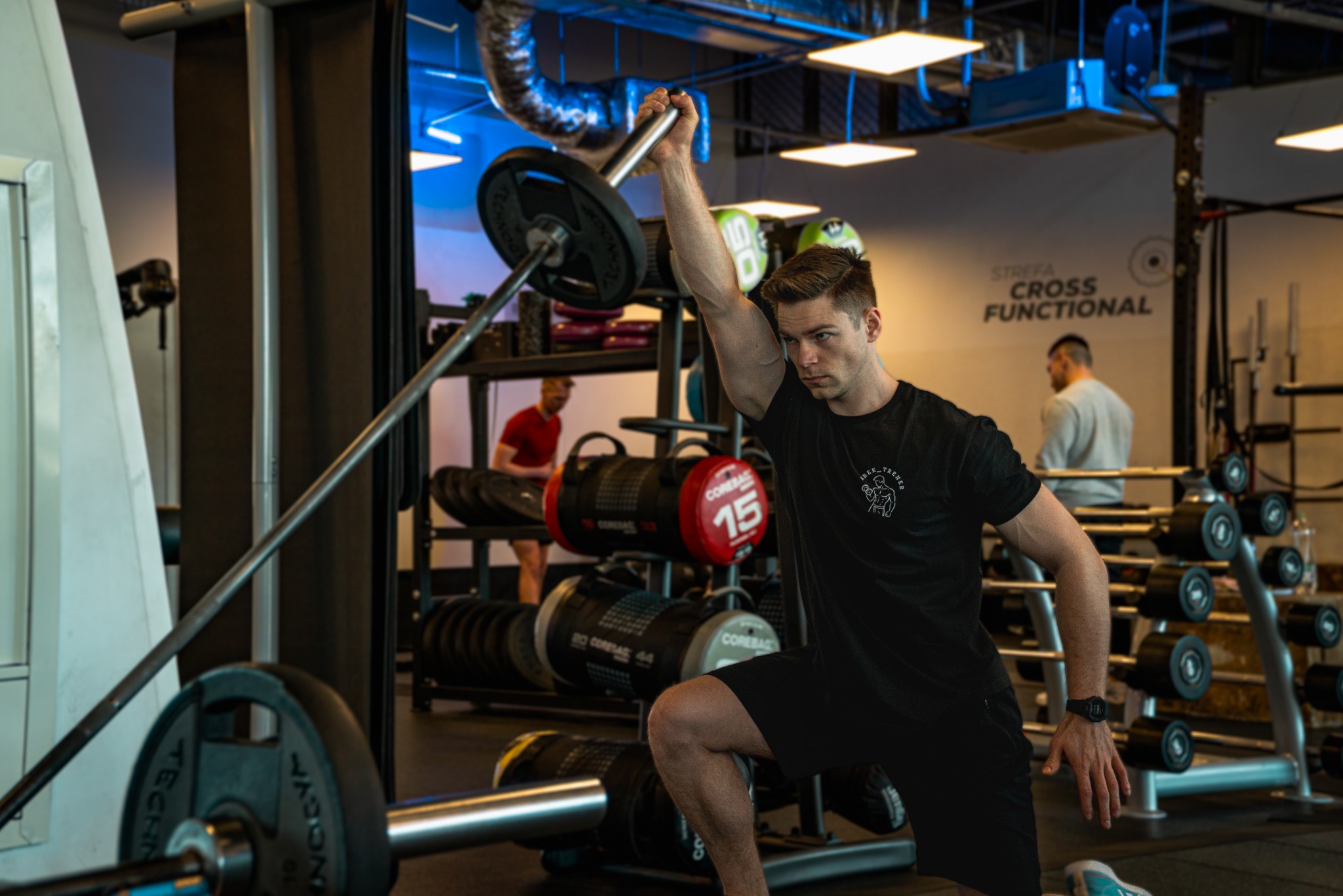 Mężczyzna w czarnym stroju sportowym klęczy w siłowni, trzymając sztangę nad głową. W tle widoczne sprzęty do ćwiczeń i napis 'Strefa Cross Functional'.