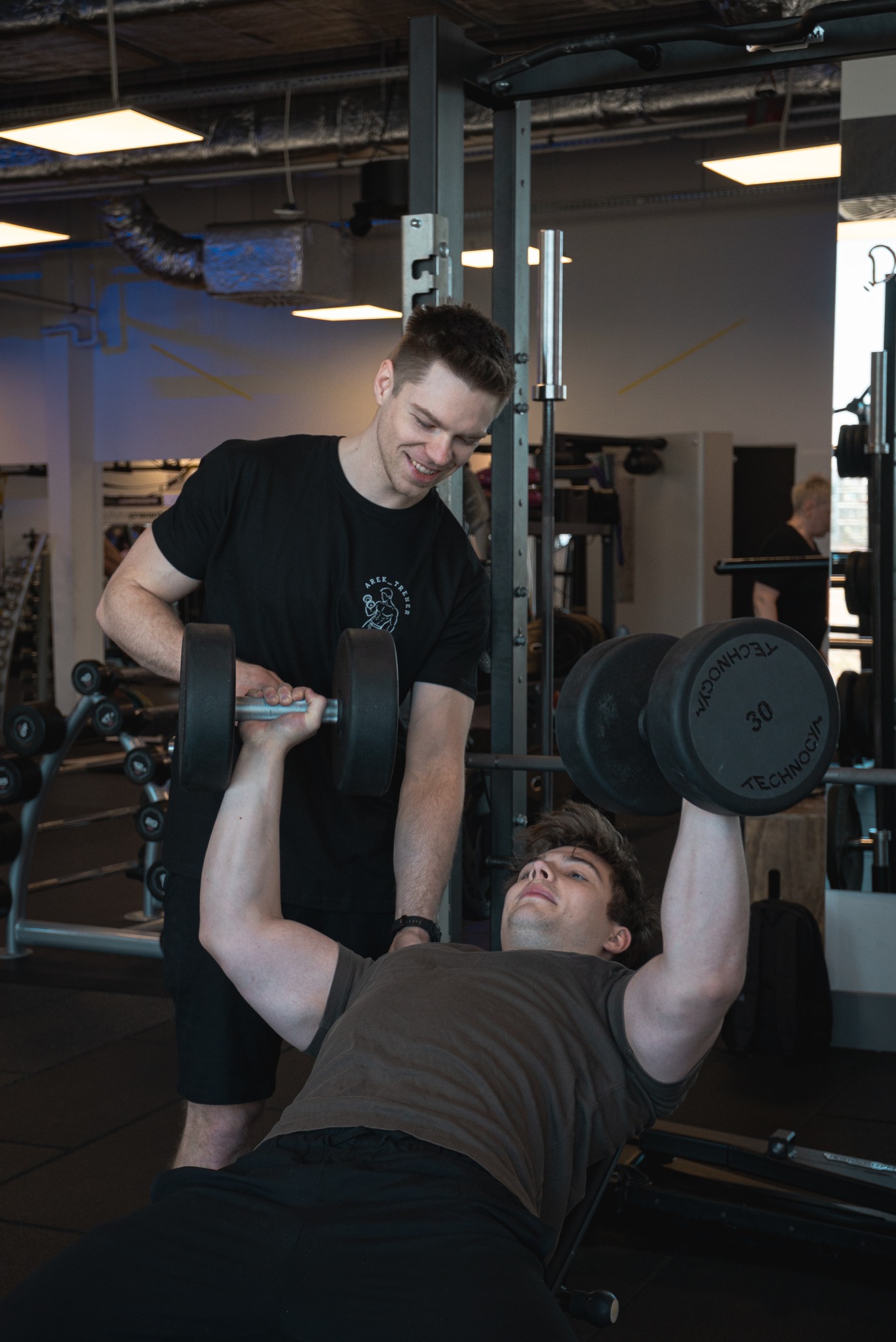 Mężczyzna ćwiczy wyciskanie sztangielek pod okiem uśmiechniętego instruktora w siłowni. Widoczne regały z obciążeniami i sprzęt do ćwiczeń.