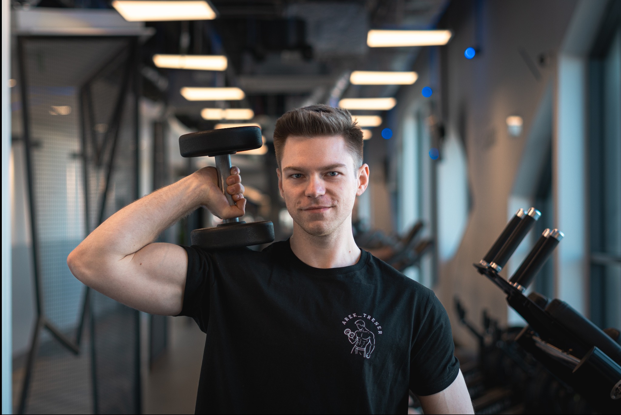 Młody mężczyzna w czarnej koszulce z logotypem, trzymający hantlę na ramieniu w nowoczesnej siłowni z niebieskimi akcentami świetlnymi.