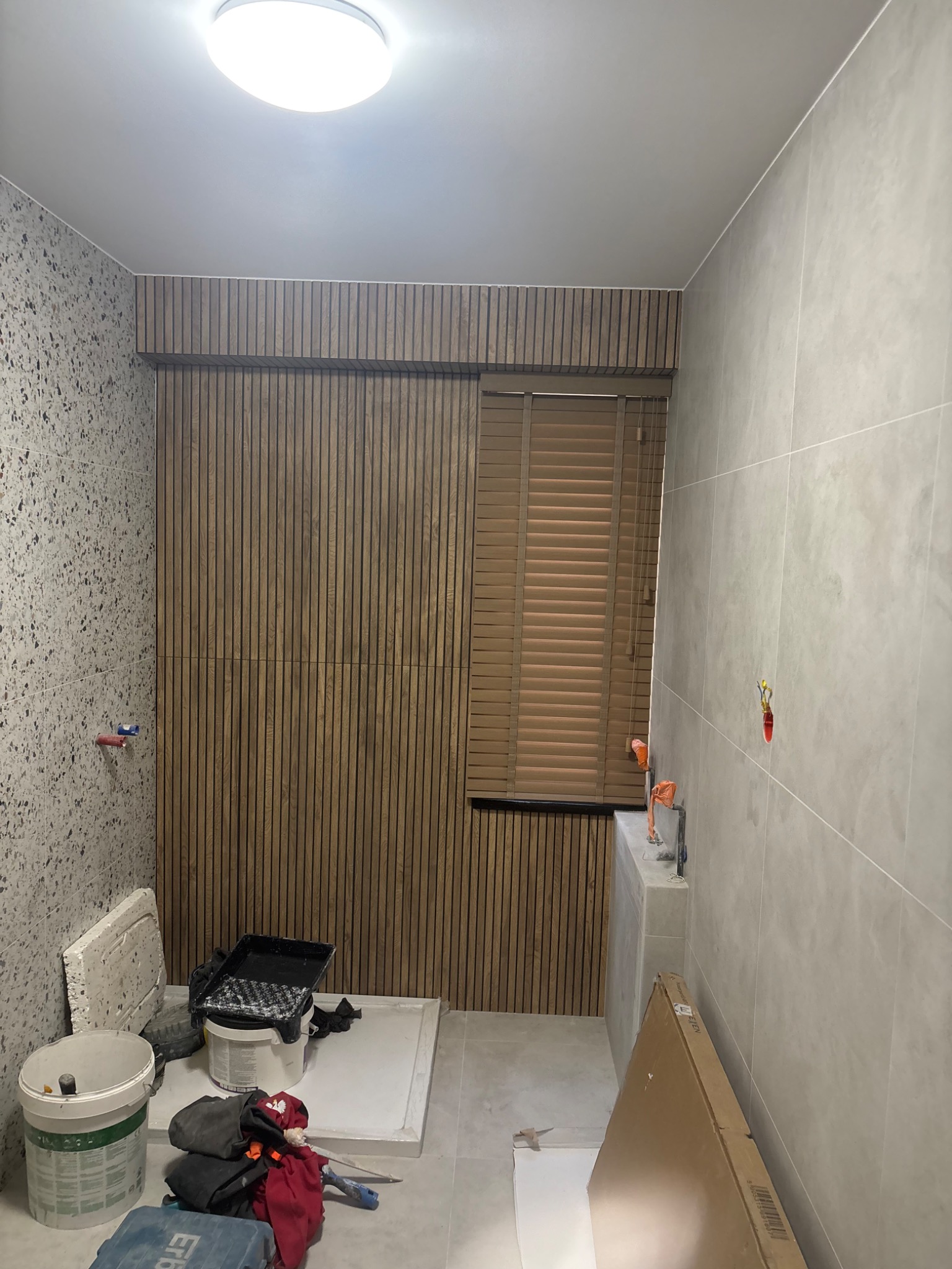 Wnętrze łazienki w trakcie prac wykończeniowych: płytki imitujące lastryko, drewniane lamele, okno z żaluzjami, widoczne narzędzia i materiały budowlane.