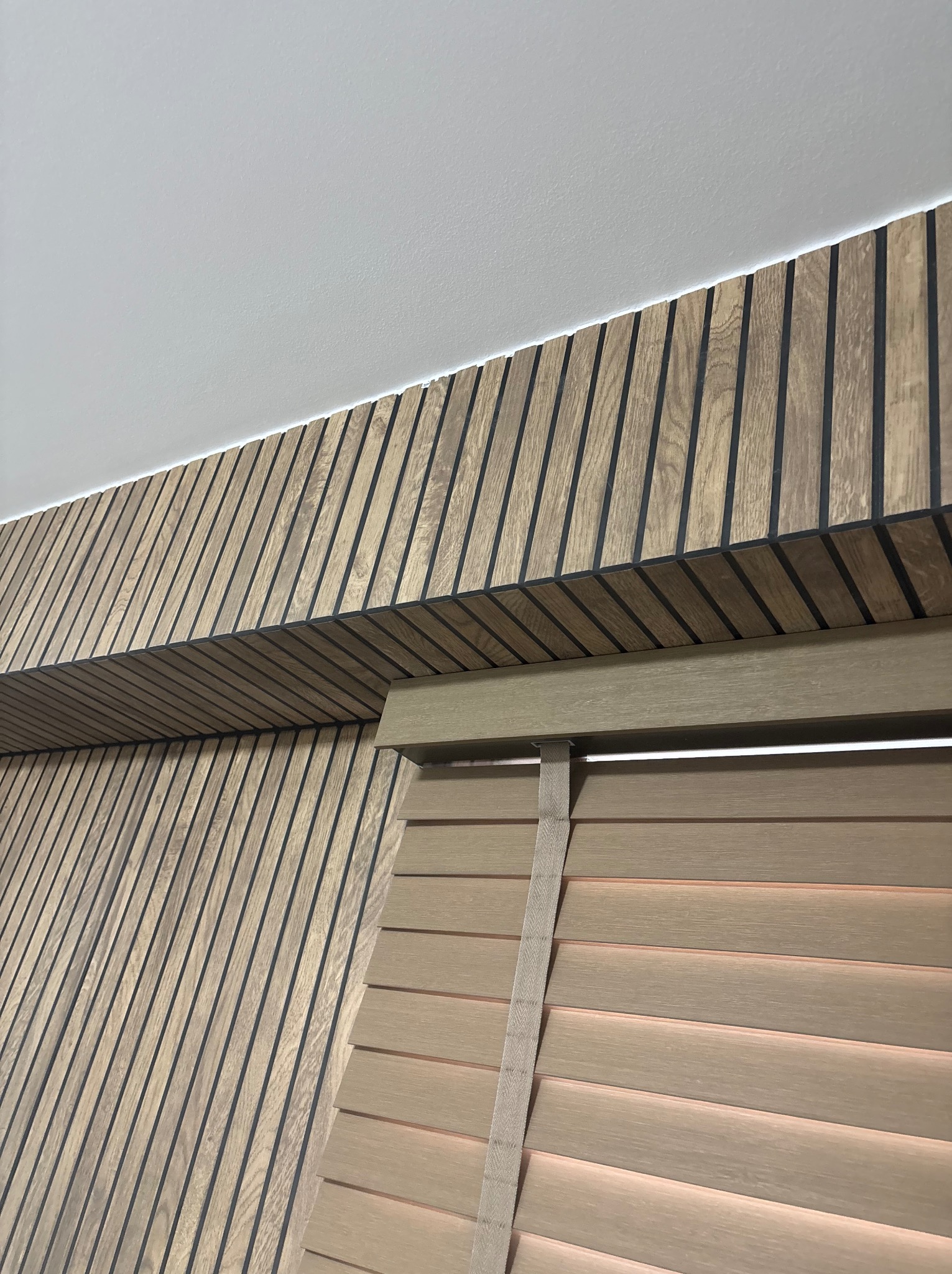 Drewniane lamele ścienne z czarnymi akcentami, połączone z drewnianymi żaluzjami w jasnym odcieniu, tworzące nowoczesny i elegancki wystrój wnętrza.