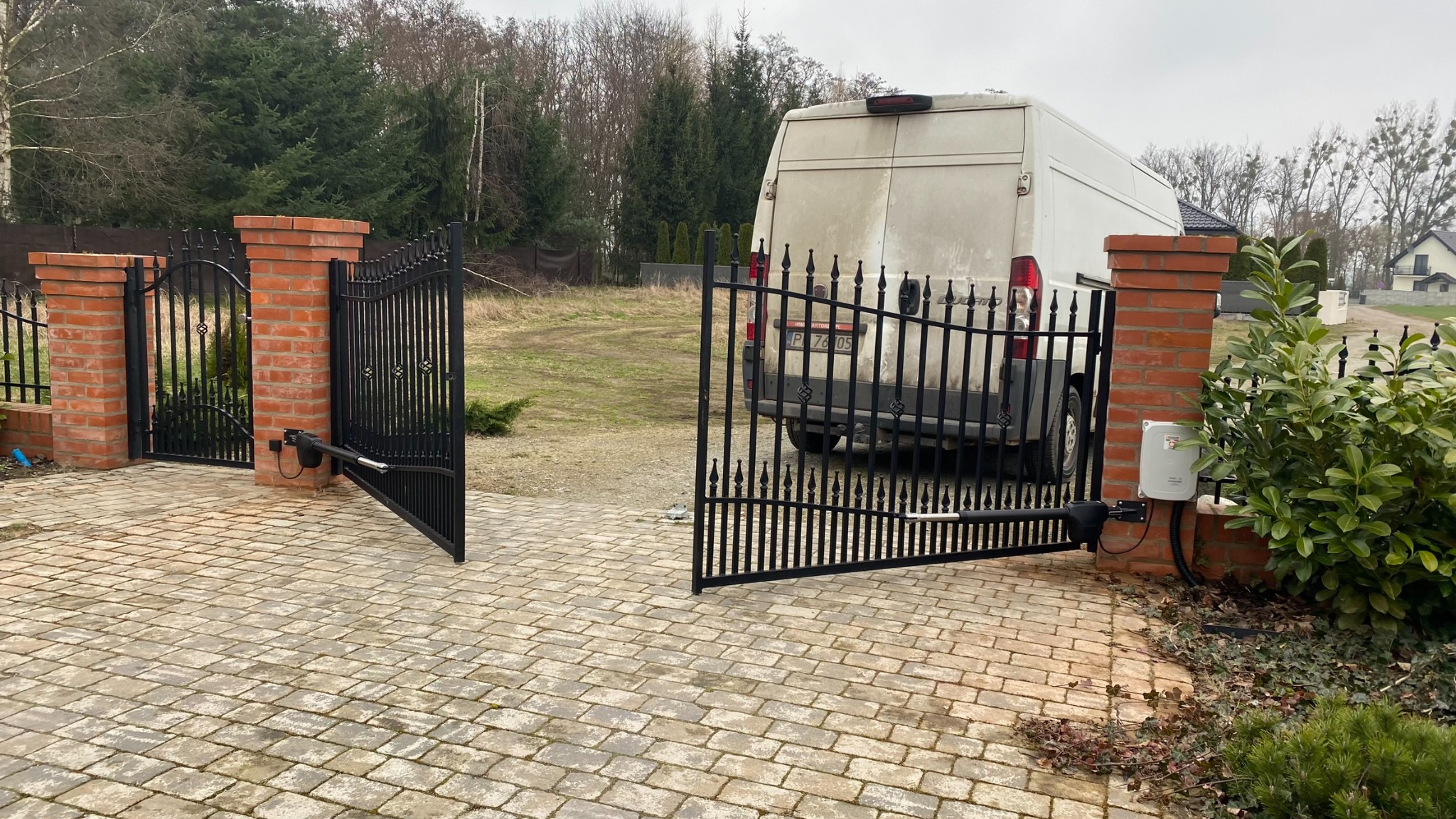 Otwarta, czarna brama wjazdowa z automatyką, zamontowana na ceglanych słupkach. W tle widoczny biały samochód dostawczy i zieleń.