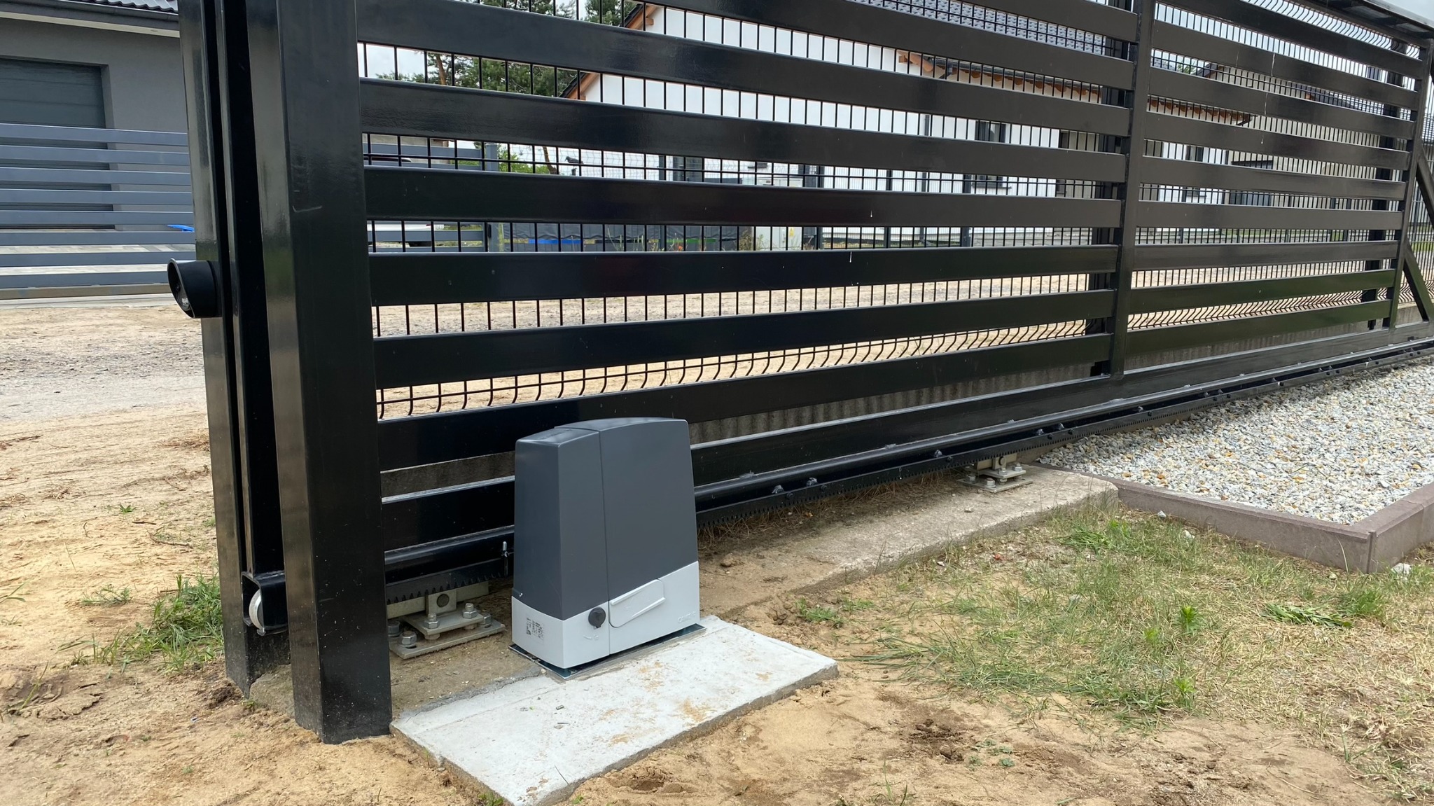 Nowoczesna, przesuwna brama wjazdowa z czarnymi, poziomymi panelami i szarym napędem automatycznym na betonowej podstawie. Widoczny fragment posesji i domu w tle.