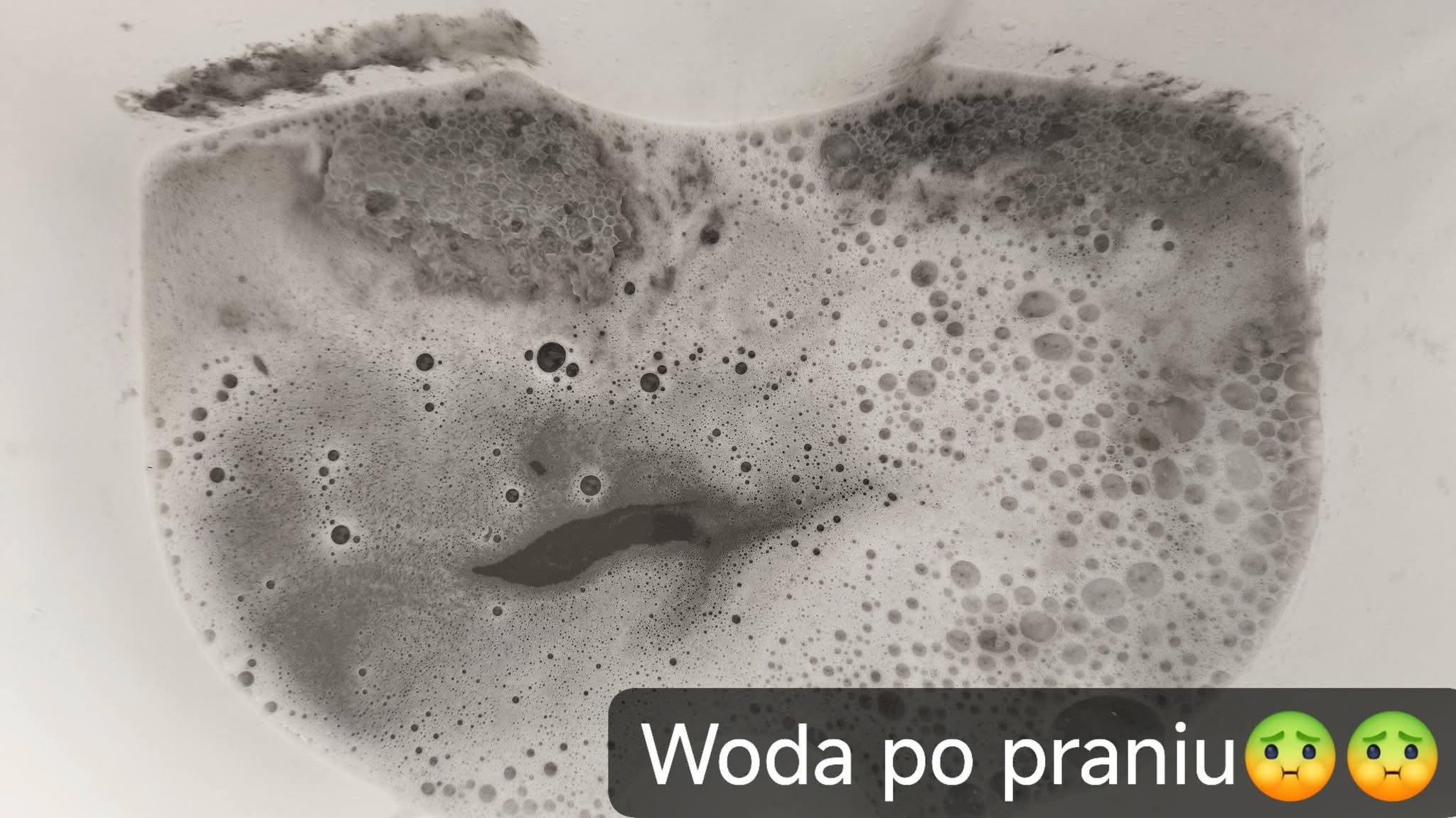 Brudna, spieniona woda po praniu tapicerki w białym wiadrze. Widoczne ciemne zanieczyszczenia i piana. Ujęcie z góry.