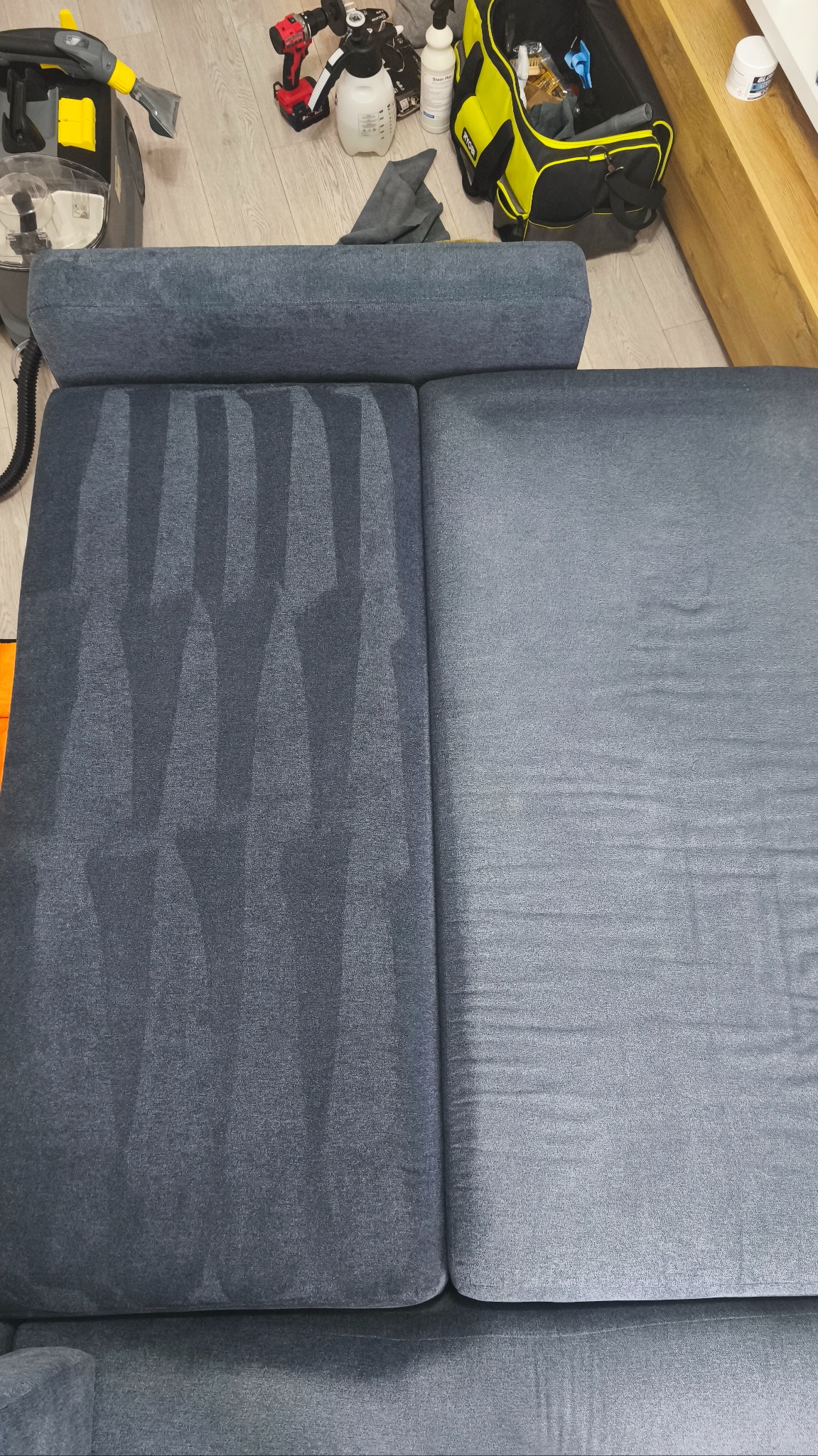 Sofa w odcieniach szarości, z widoczną różnicą po praniu na jednej części. Obok sprzęt do czyszczenia i narzędzia. Widok z góry, ukazujący efekt czyszczenia.