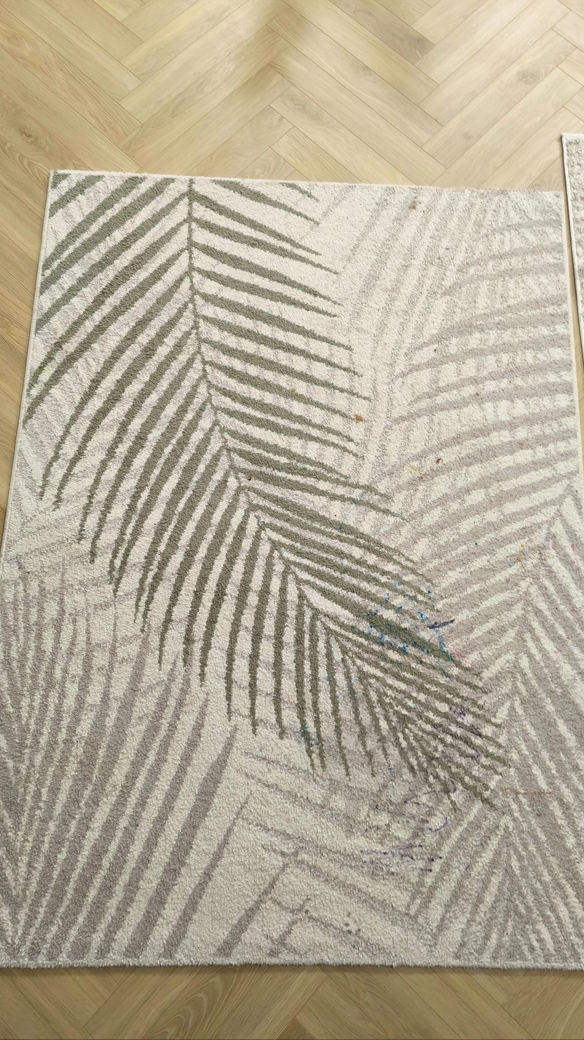 Jasny dywan z motywem liści palmowych na parkiecie. Widoczne plamy i zabrudzenia na powierzchni dywanu, sugerujące potrzebę czyszczenia.