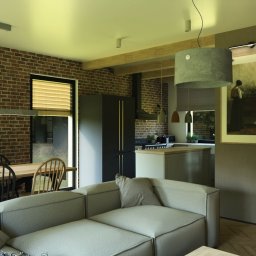 Lofera - Wnętrze domu z ceglaną ścianą, drewnianym stołem i szarą sofą. Widoczna kuchnia z wyspą i schody. Nad stołem podłużna lampa, nad wyspą okrągła.