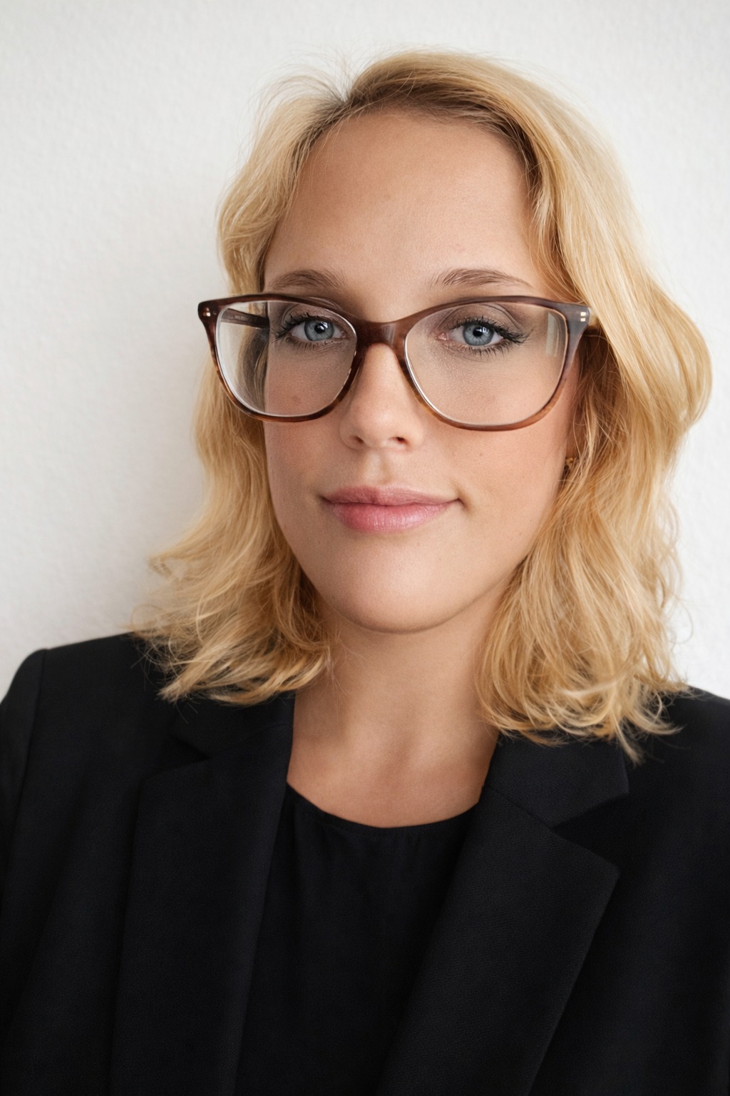 Portret kobiety w okularach i czarnej marynarce na jasnym tle. Uśmiechnięta, z delikatnym makijażem, patrzy wprost. Profesjonalny wizerunek.