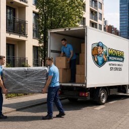 Movers Przeprowadzki - Transport Katowice i okolice - Pracownicy w niebieskich koszulkach wnoszą zapakowany mebel do białej furgonetki z logo firmy przeprowadzkowej na tle nowoczesnego budynku w Katowicach.
