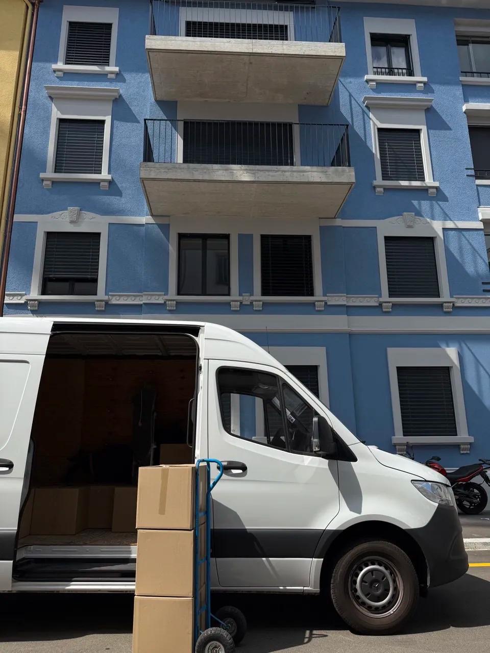 Otwarty biały furgon z kartonami na wózku transportowym na tle błękitnego budynku z balkonami. Widoczny motocykl w tle. Słoneczny dzień.