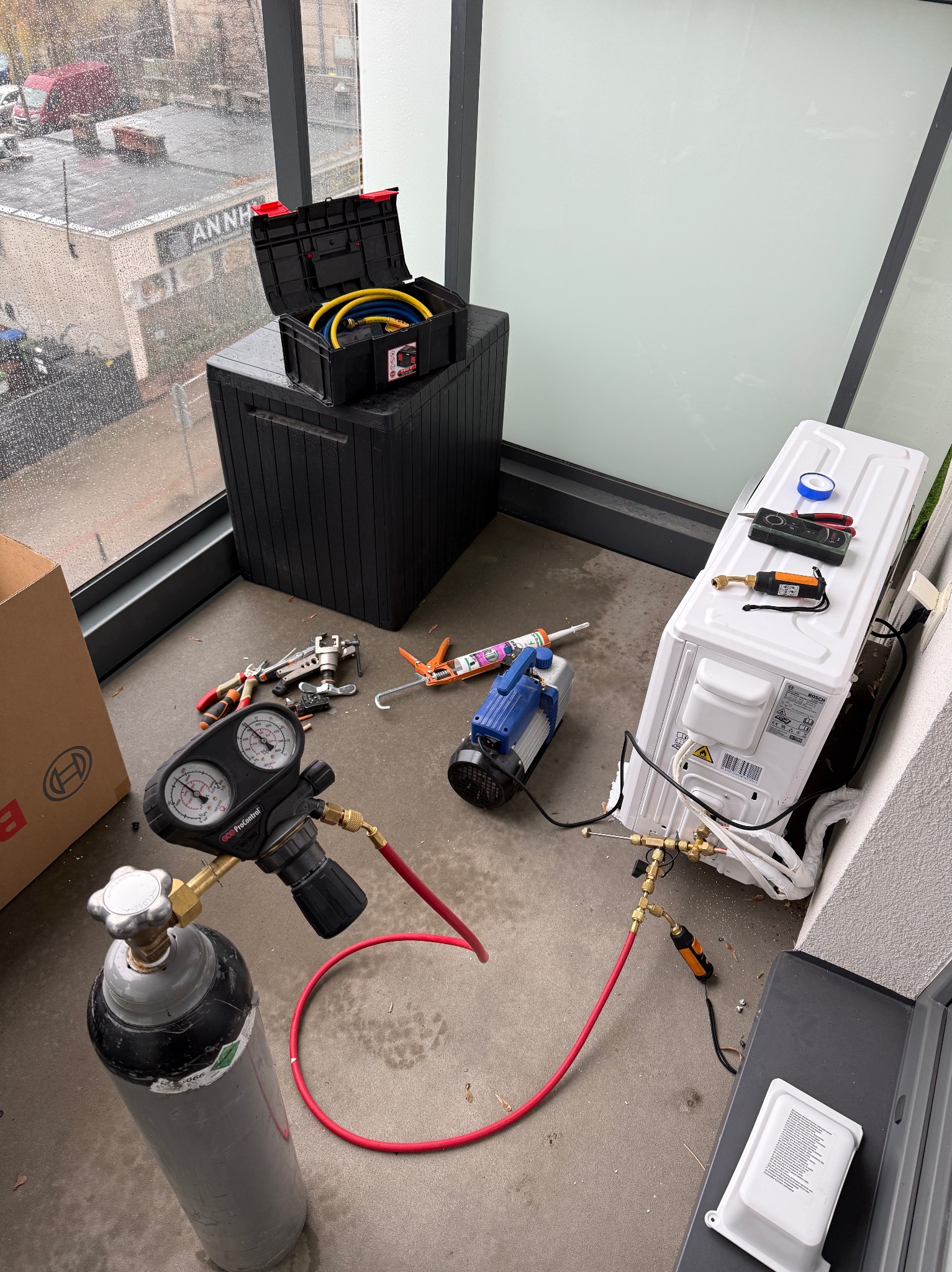 Montaż klimatyzacji na balkonie: butla z gazem, pompa próżniowa i narzędzia obok jednostki zewnętrznej. Widok z góry na balkon z widokiem na miasto w tle.