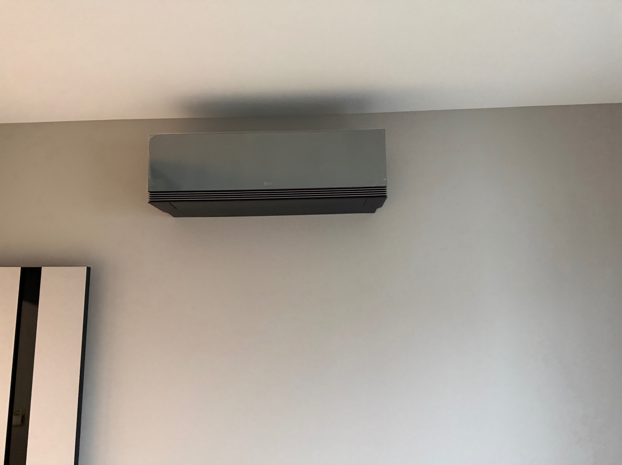 Szary klimatyzator LG zamontowany na jasnej ścianie w pomieszczeniu. Widoczny fragment szafki z czarno-białymi pasami. Minimalistyczny design.