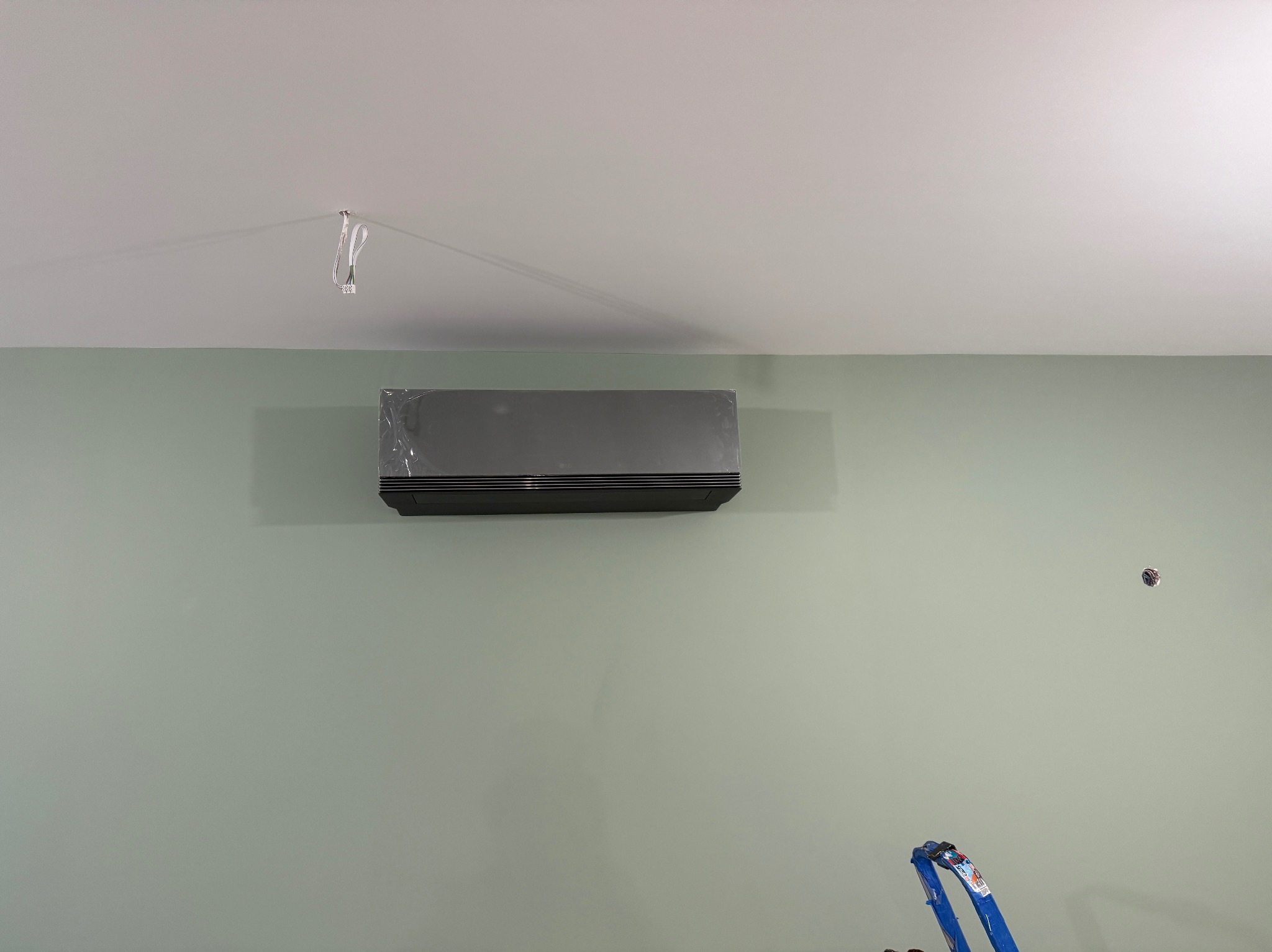 Szary klimatyzator ścienny, jeszcze w folii ochronnej, zamontowany na zielonej ścianie. Widoczne przewody elektryczne i fragment drabiny.