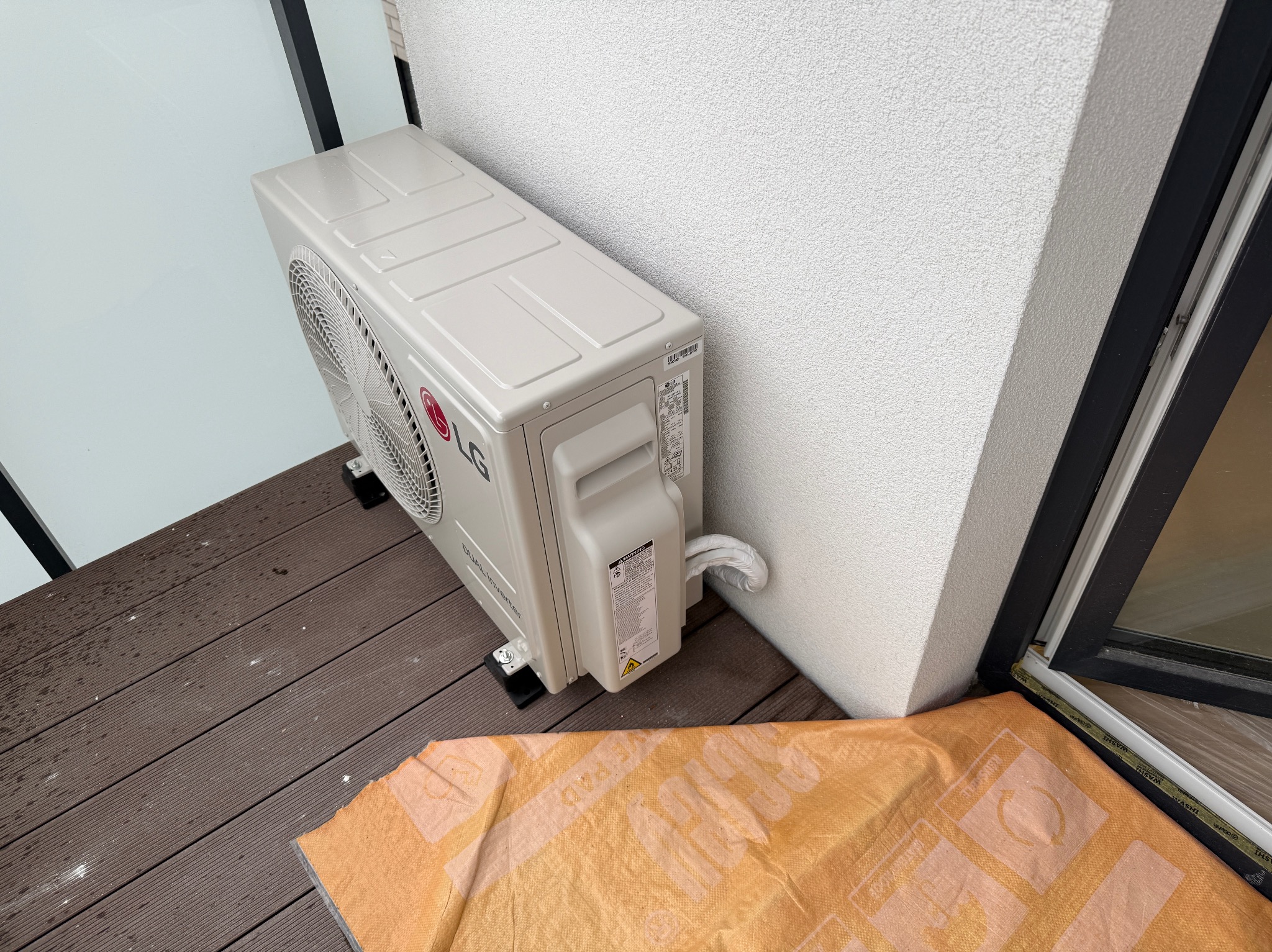 Zewnętrzna jednostka klimatyzacji LG Dual Inverter zamontowana na balkonie z drewnianą podłogą, zabezpieczona pomarańczową folią ochronną.