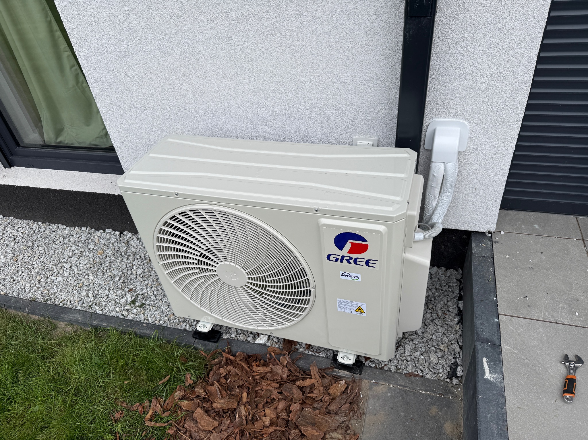 Zewnętrzna jednostka klimatyzacji Gree, zamontowana przy białej ścianie domu, otoczona kamieniami i korą. Obok leży klucz nastawny.