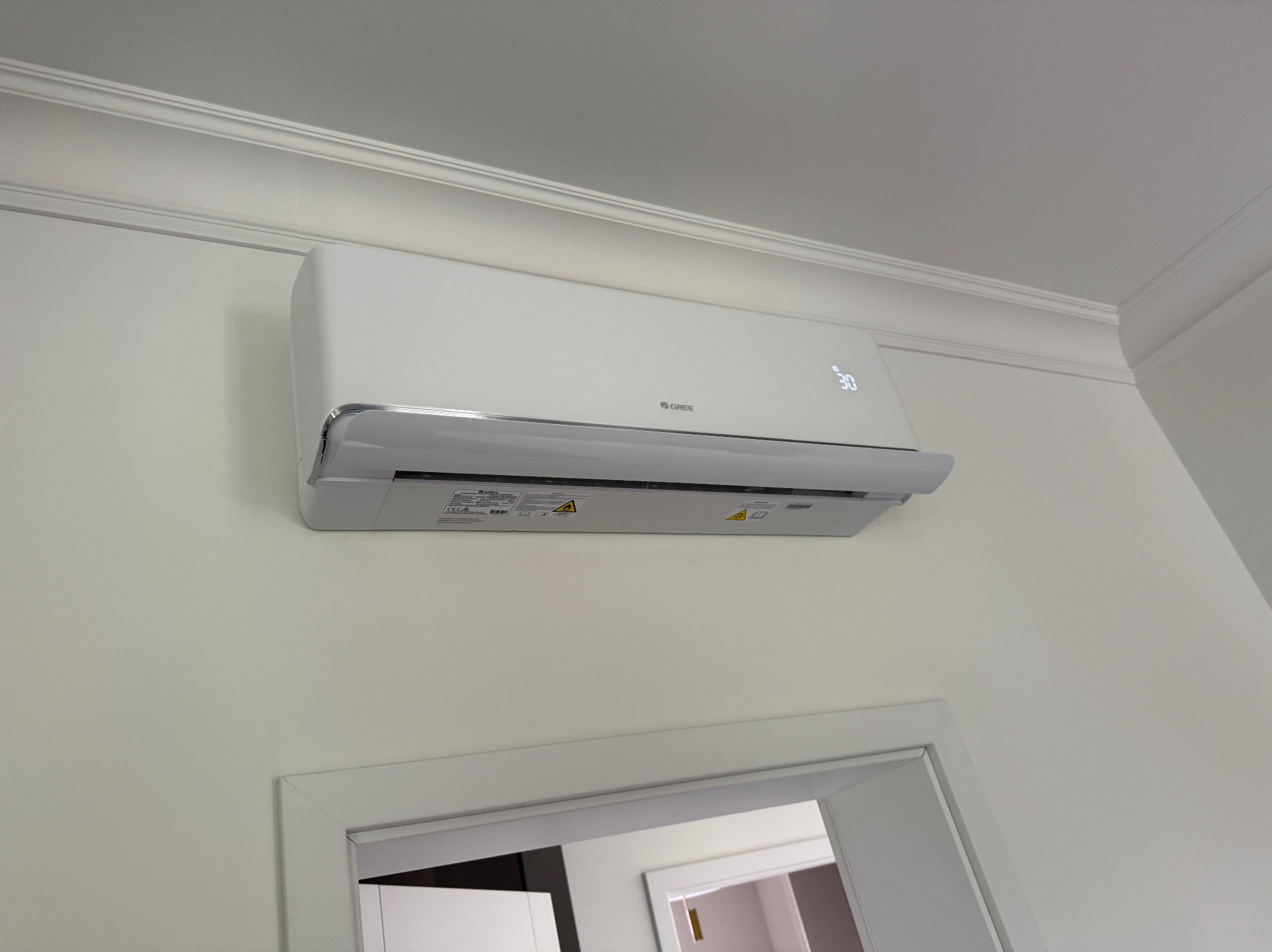 Biała klimatyzacja Gree zamontowana na ścianie w jasnym wnętrzu, widoczny fragment sufitu z ozdobną listwą i górna część drzwi. Minimalistyczny design.
