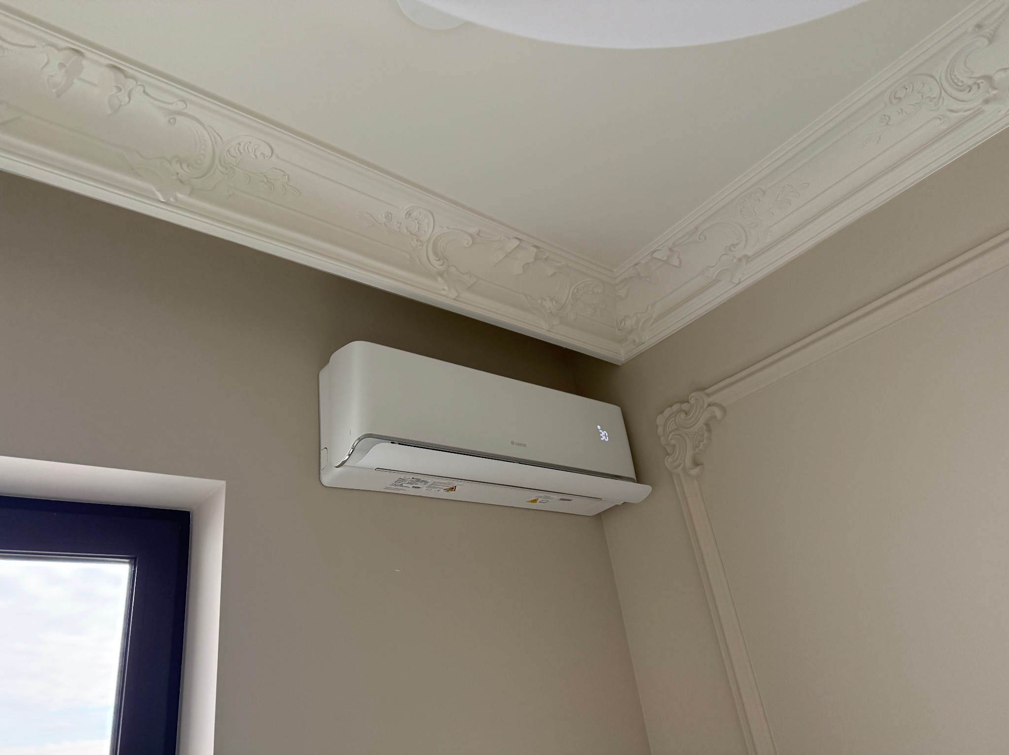 Biała klimatyzacja na ścianie w pokoju z ozdobnym sufitem i ramą okienną. Widoczny wyświetlacz temperatury na urządzeniu.