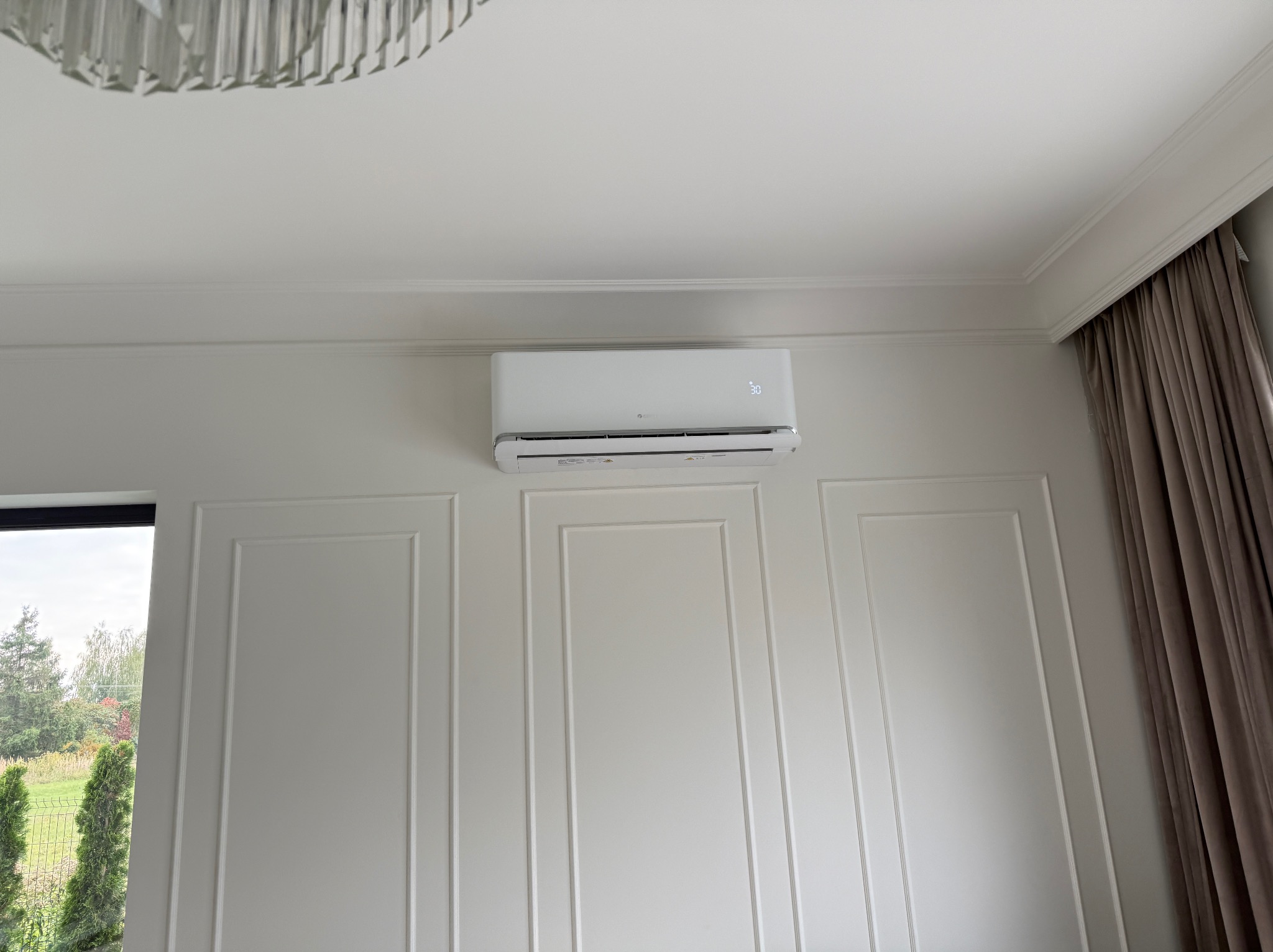 Biały klimatyzator na ścianie z dekoracyjnymi panelami w jasnym wnętrzu. Widoczne okno z zielenią na zewnątrz i fragment zasłony. Nowoczesny design.