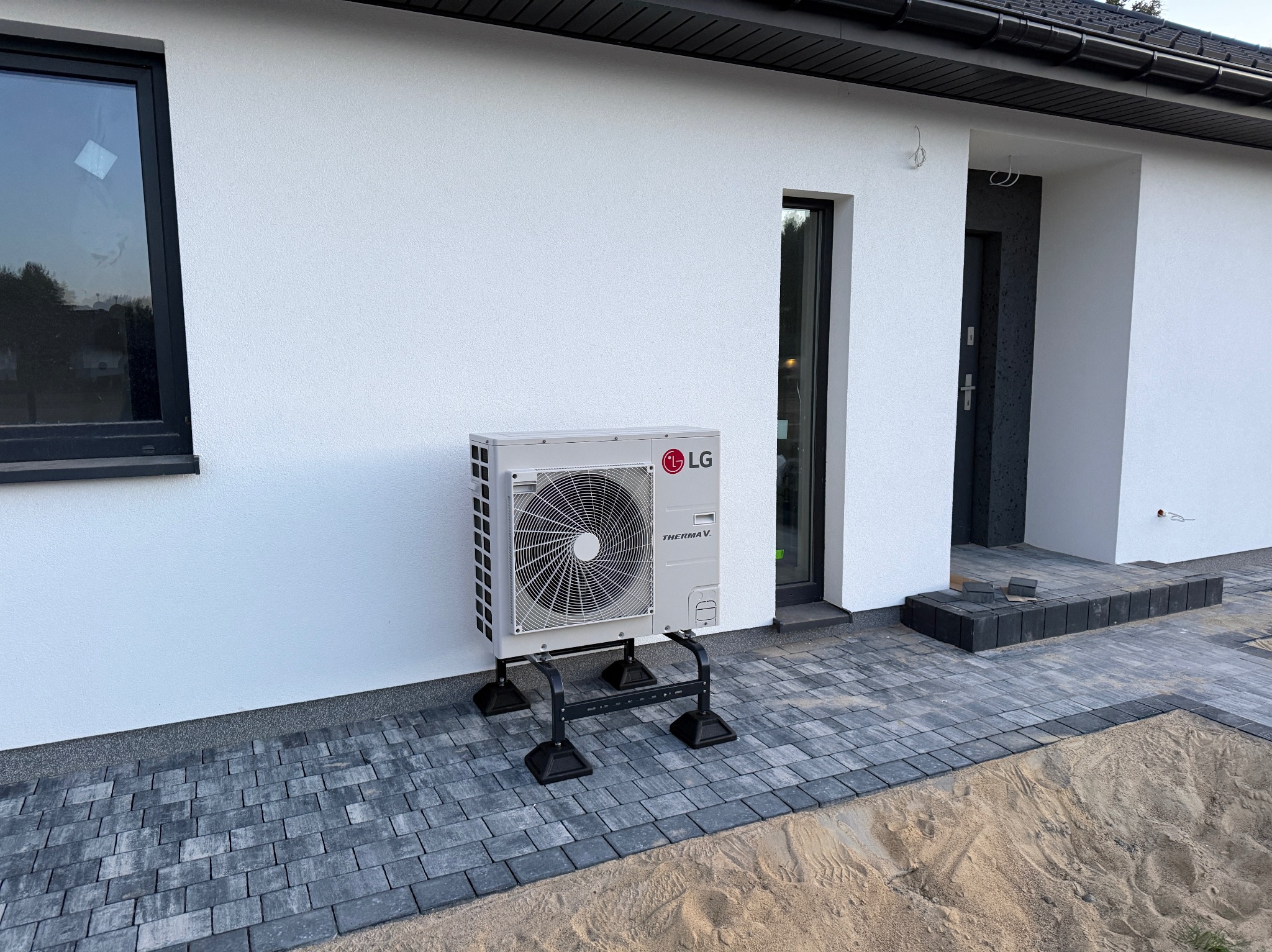 Biała jednostka zewnętrzna klimatyzacji LG Therma V na czarnym stelażu, zamontowana przy białej ścianie domu z szarą kostką brukową i piaskiem w tle.