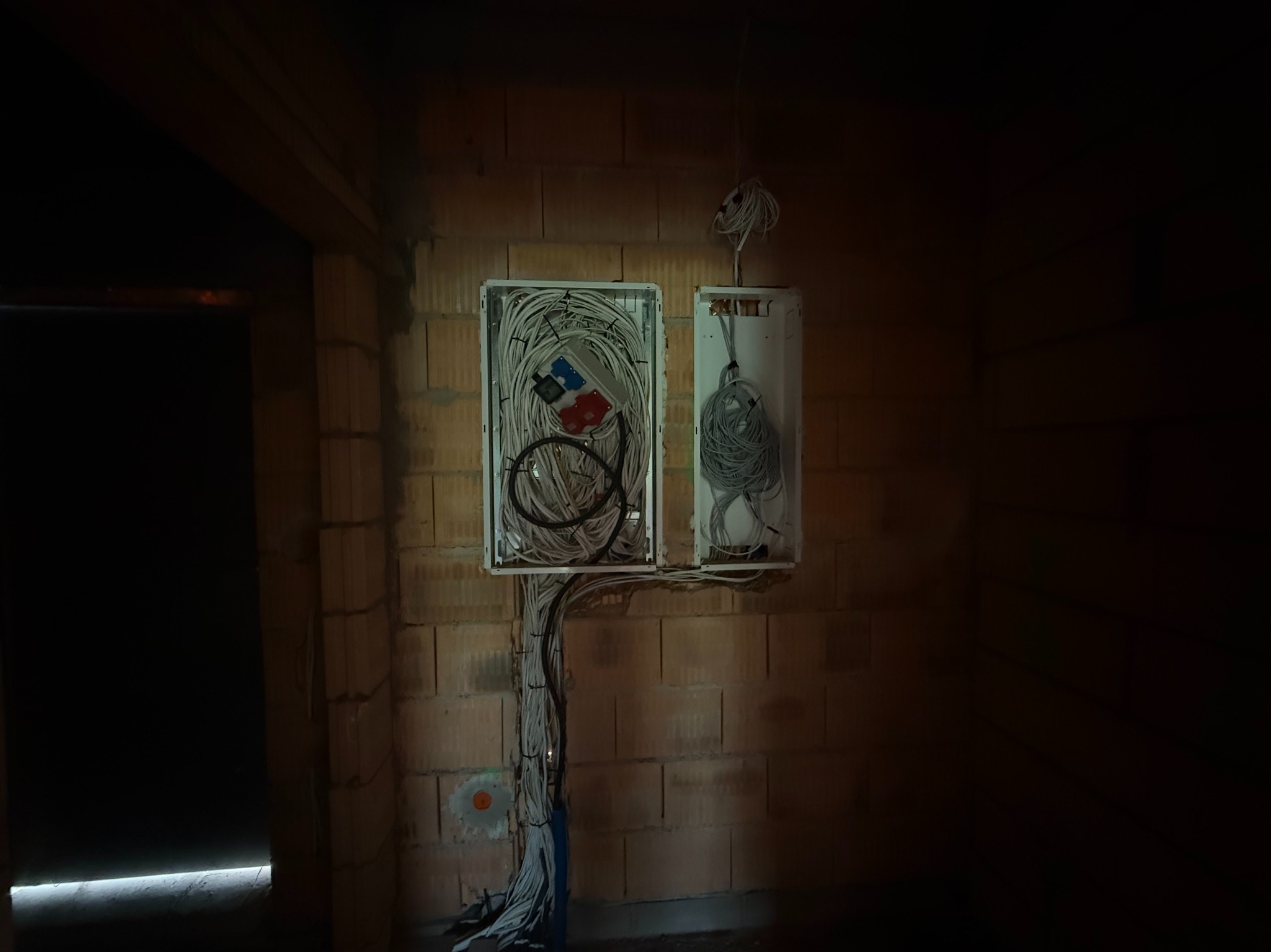 Rozdzielnia elektryczna z licznymi przewodami na ceglanej ścianie w surowym wnętrzu. Widoczne kable w białych korytkach i puszka podtynkowa.