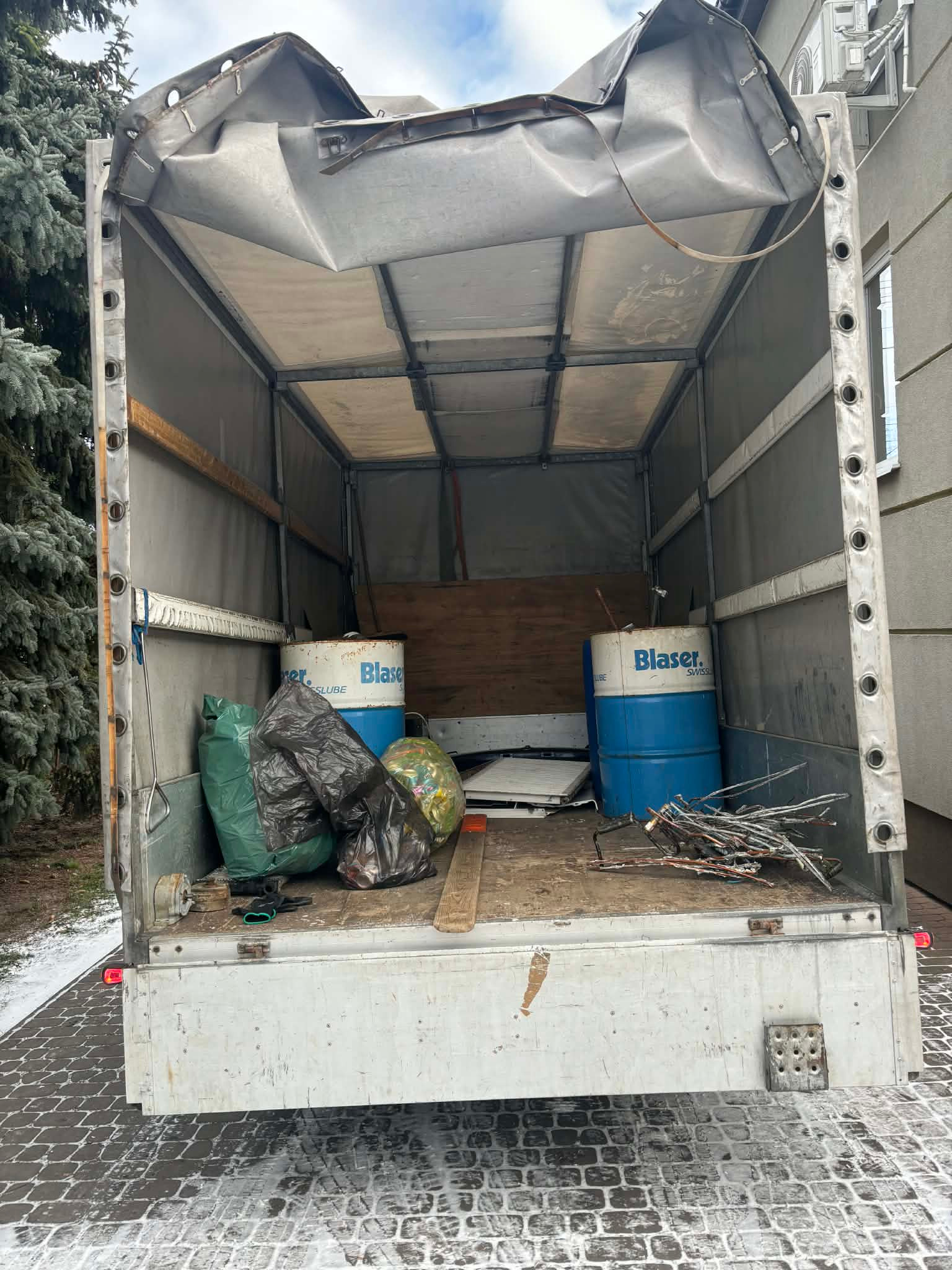 Otwarta paka busa z workami, beczkami i odpadami. Widoczne elementy budynku w tle. Ujęcie z tyłu pojazdu na kostce brukowej.