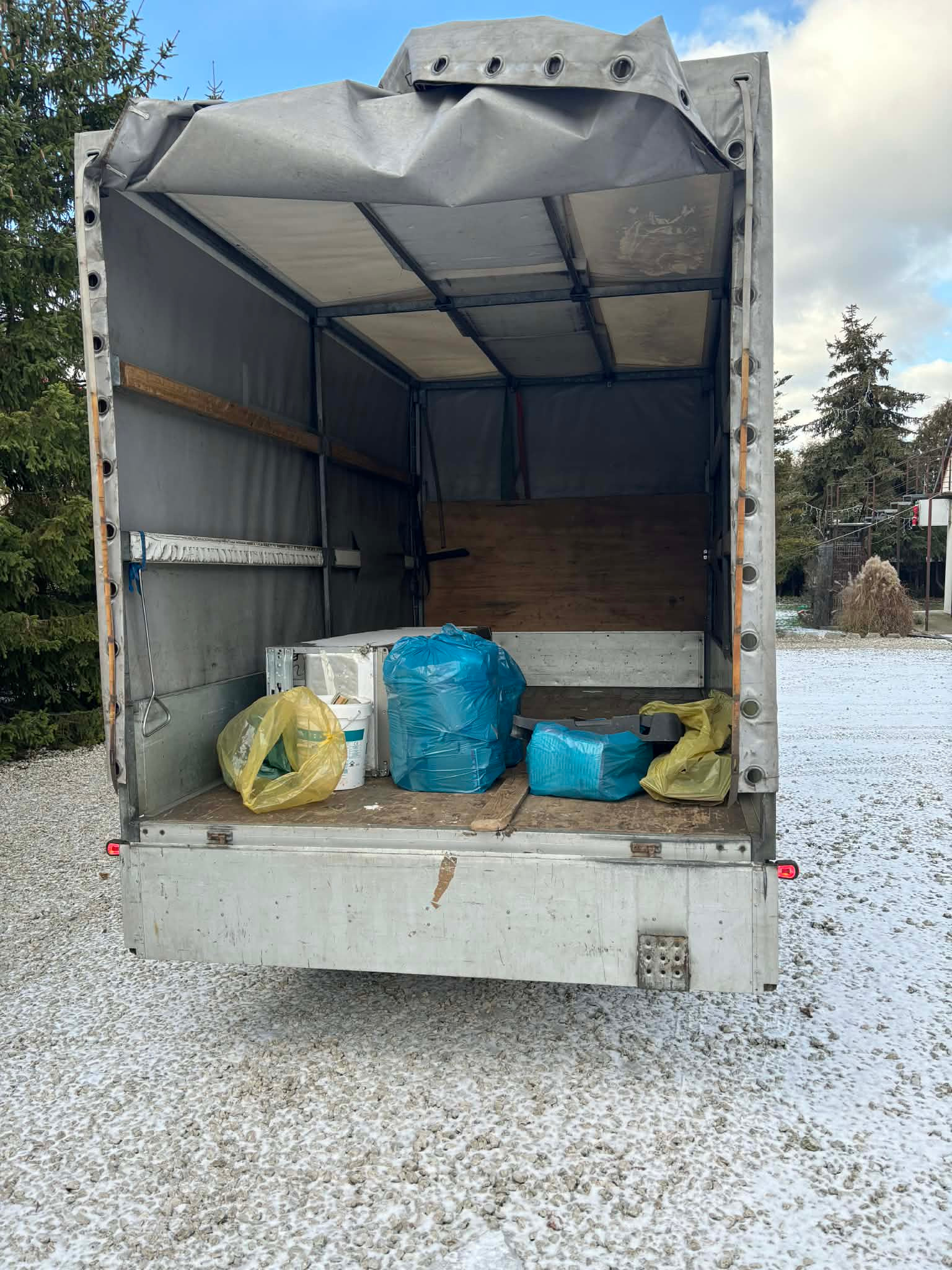 Otwarta paka busa z workami i wiadrami, prawdopodobnie po opróżnianiu mieszkania. Zimowy krajobraz w tle, śnieg na ziemi. Widok z tyłu pojazdu.
