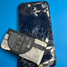 iMate Mateusz Płacheta - Rozbity iPhone z odłączoną baterią na niebieskiej macie serwisowej. Widoczne pęknięcia na obudowie i bateria z oznaczeniami producenta. Uszkodzony smartfon po demontażu.