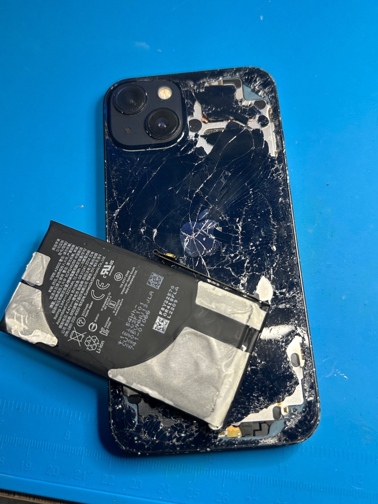 Rozbity iPhone z odłączoną baterią na niebieskiej macie serwisowej. Widoczne pęknięcia na obudowie i bateria z oznaczeniami producenta. Uszkodzony smartfon po demontażu.