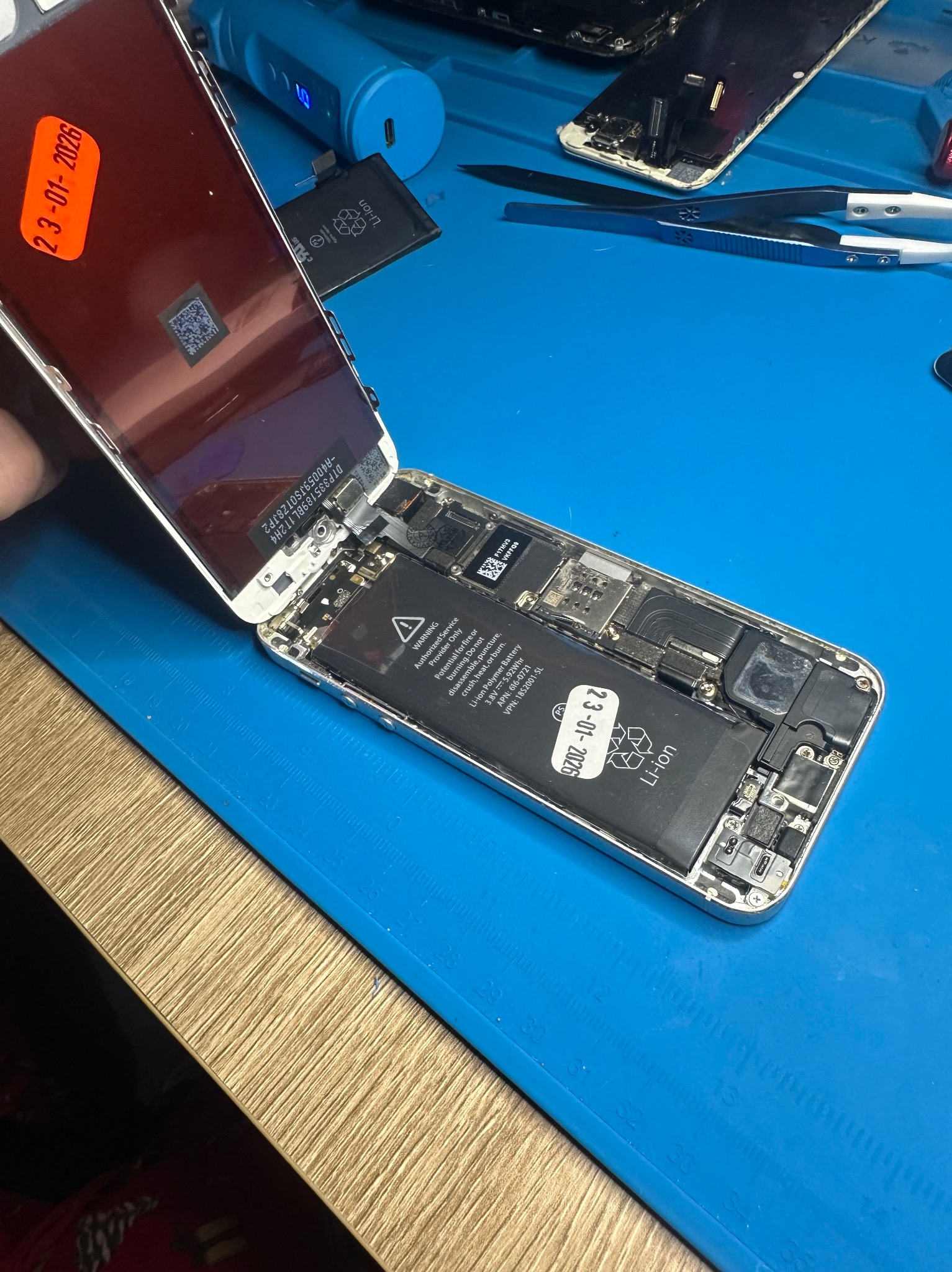 Rozłożony iPhone na niebieskiej macie serwisowej, widoczna bateria z datą ważności 23-01-2026. Ekran odchylony, ukazujący wnętrze urządzenia. Narzędzia serwisowe w tle.