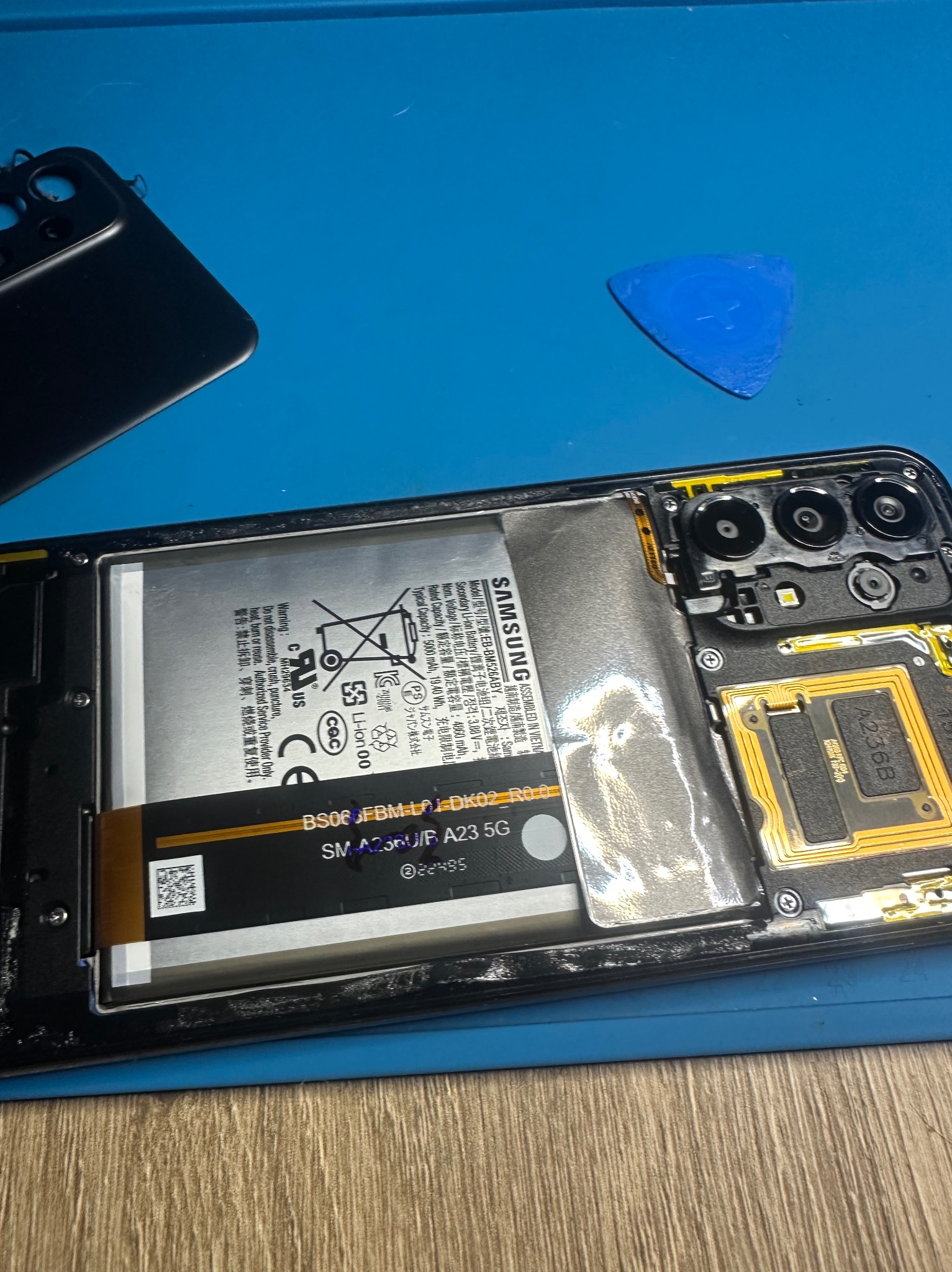 Otwarty smartfon Samsung A23 5G z widoczną baterią, elementami elektroniki i narzędziem do otwierania obudowy na niebieskiej macie serwisowej.