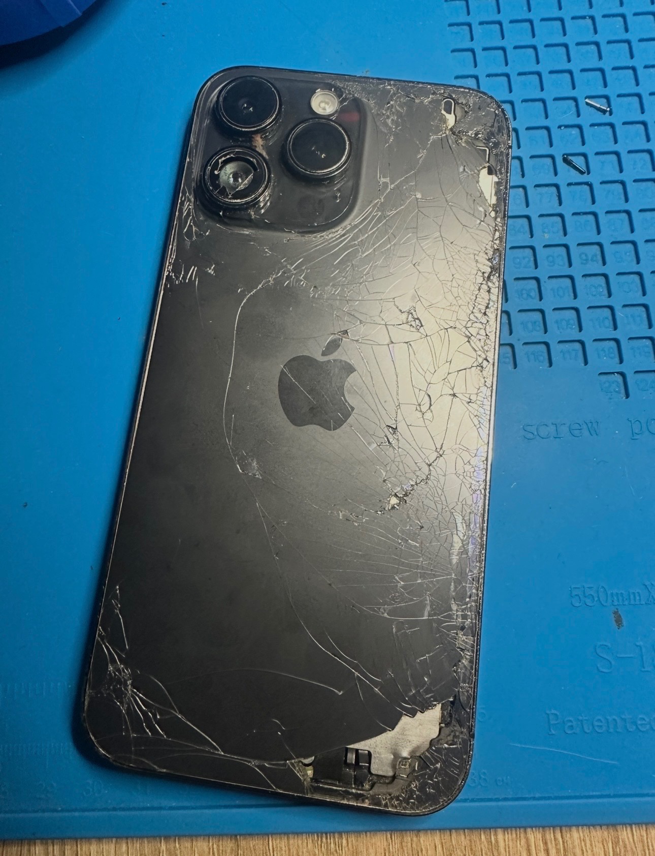 Rozbity iPhone z widocznymi pęknięciami na obudowie i uszkodzonymi soczewkami aparatu leży na niebieskiej macie serwisowej. Widoczne ślady naprawy.