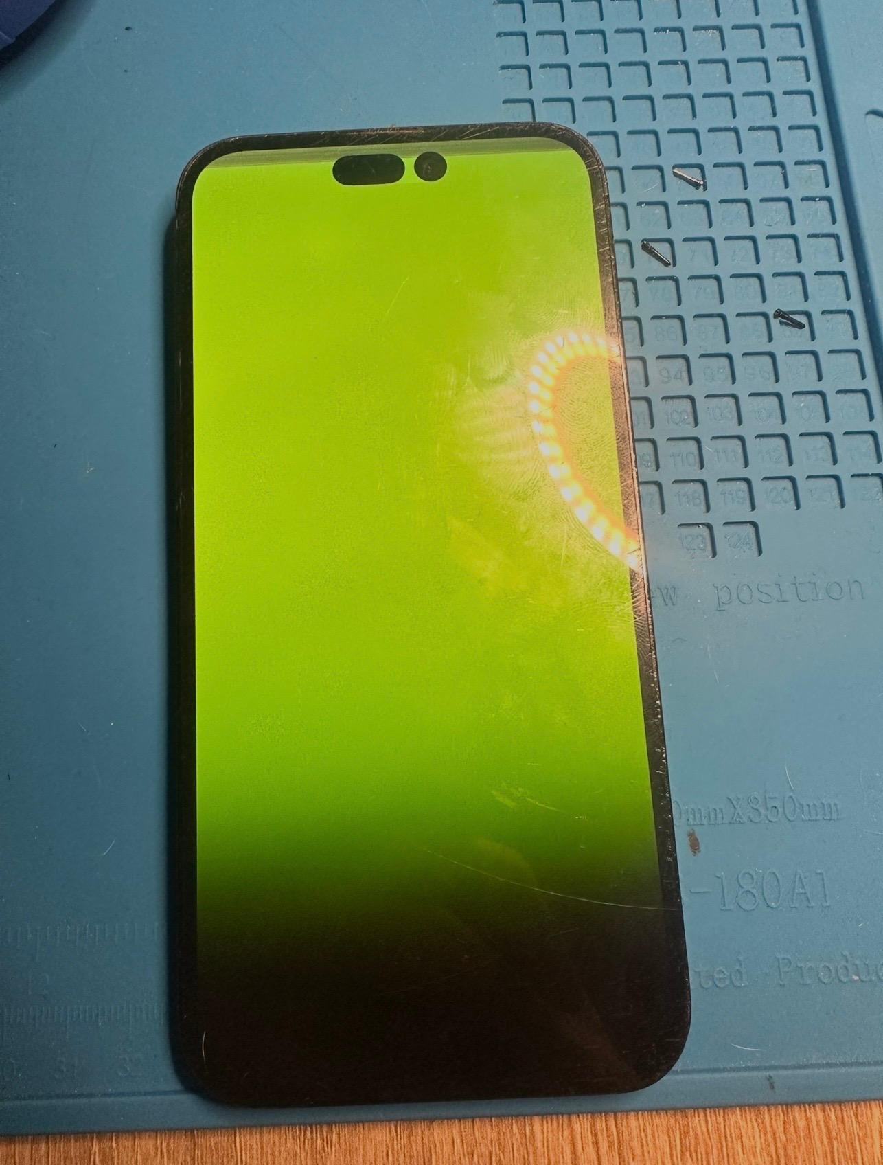 Uszkodzony smartfon z zielonym ekranem leżący na macie serwisowej, obok widoczne drobne śrubki. Awaria ekranu telefonu.