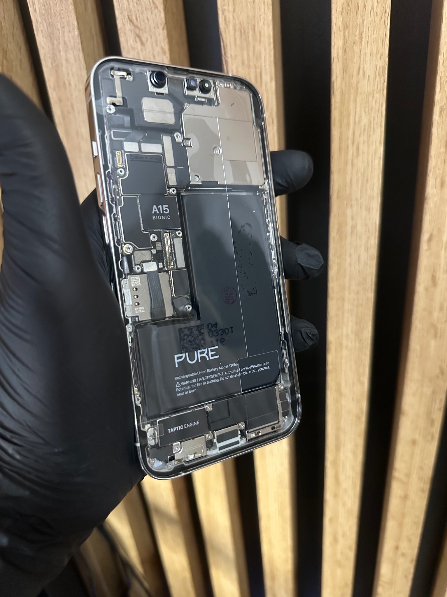 Rozłożony iPhone 14 z widoczną baterią PURE i chipem A15 Bionic, trzymany w dłoni w czarnej rękawiczce na tle drewnianej ściany. Detaliczna prezentacja wnętrza urządzenia.